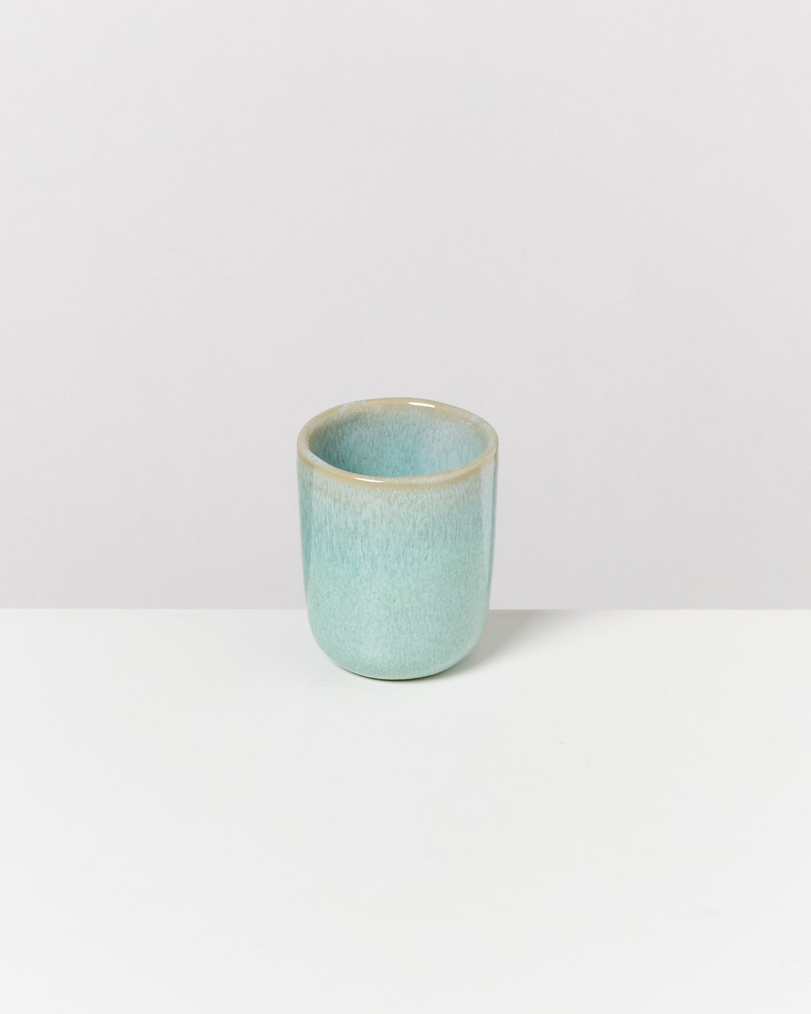 Ein kleiner, hellblauer Keramikbecher steht auf einem weißen Untergrund vor einem grauen Hintergrund.