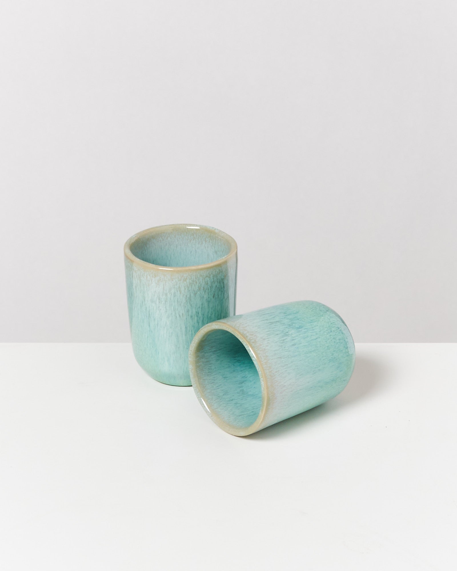 Zwei hellblaue Keramikbecher, einer steht aufrecht, der andere liegt auf der Seite, auf einem weißen Hintergrund.