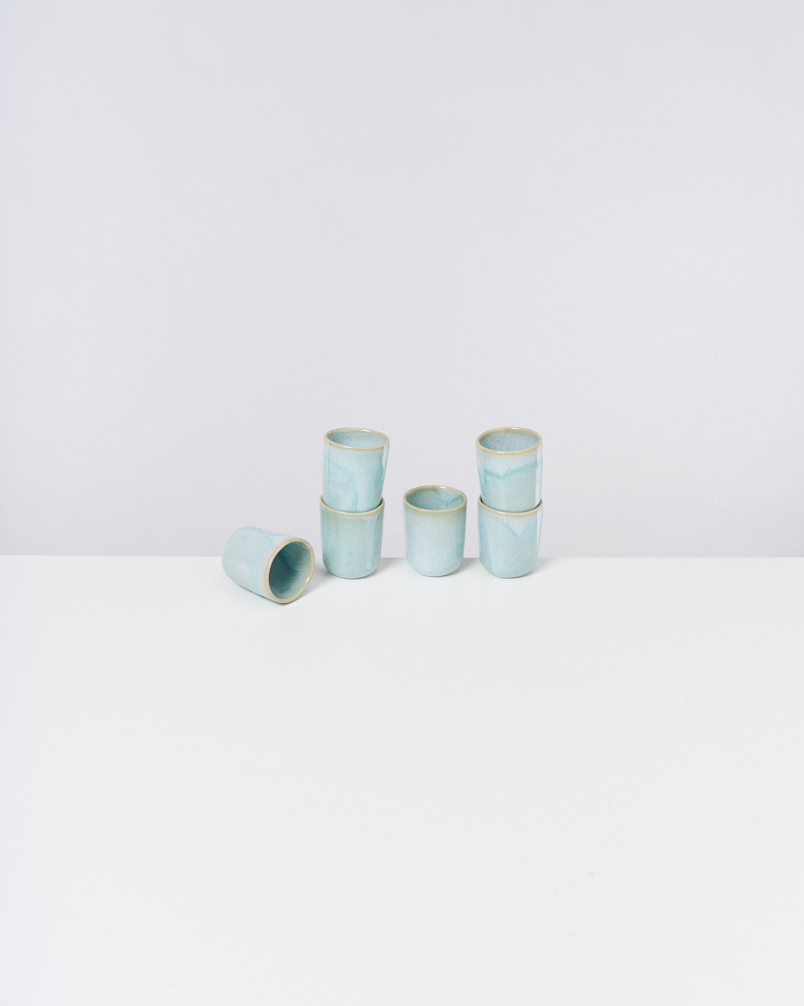 Sechs hellblaue Keramiktassen stehen auf einem weißen Tisch, eine liegt seitlich davor.