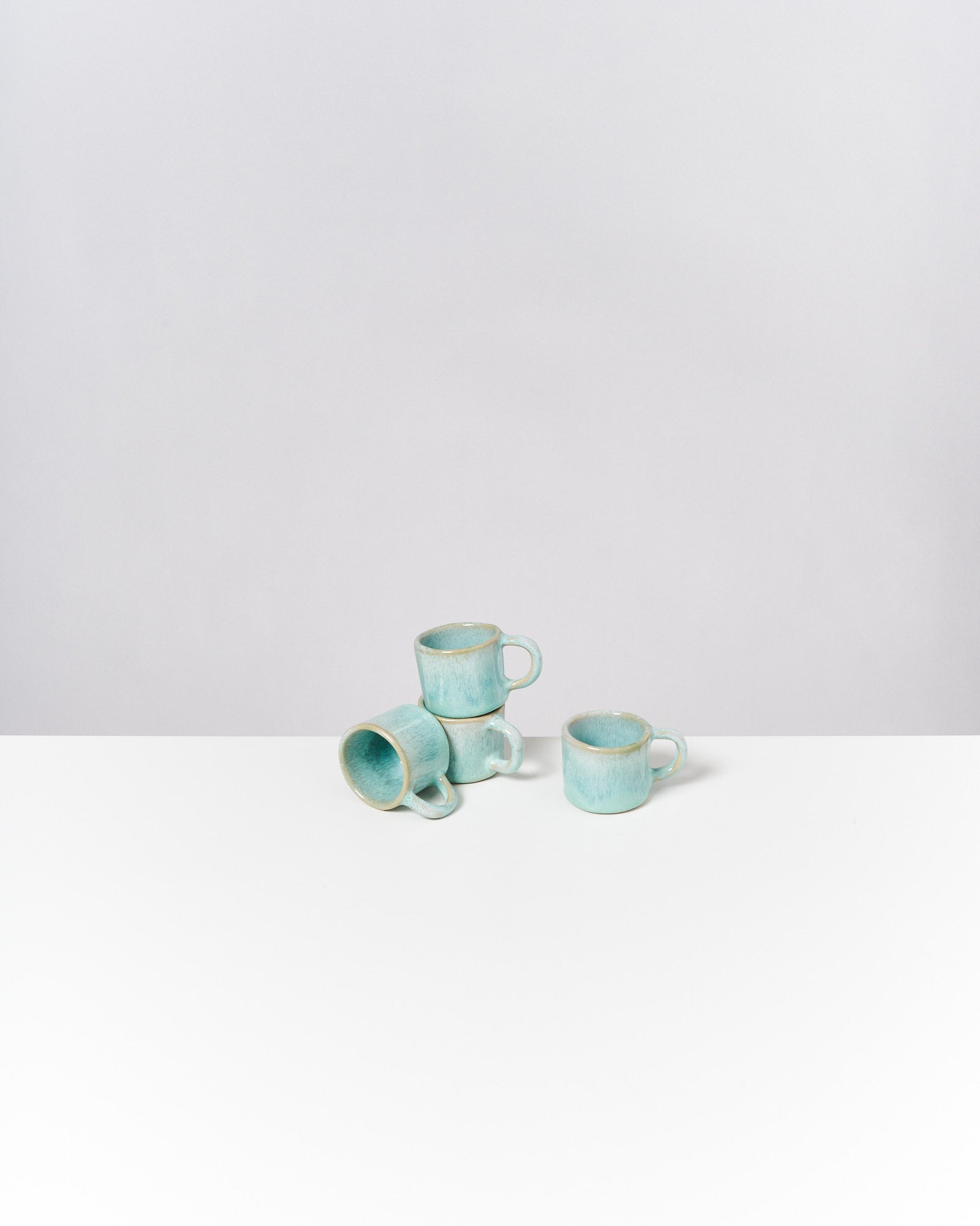 Vier hellblaue Tassen stehen ineinander gestapelt auf einem weißen Tisch vor einem grauen Hintergrund.