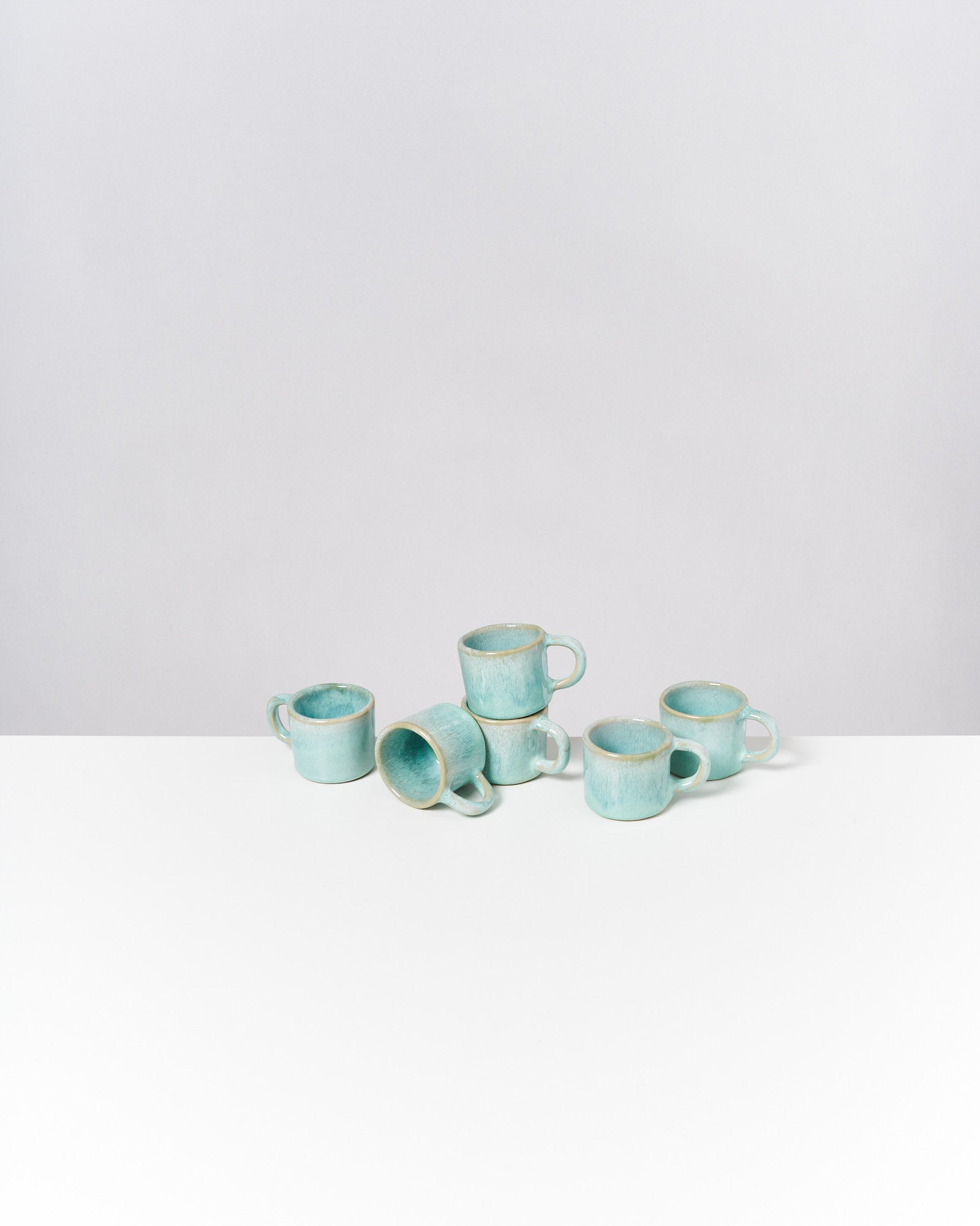 Sechs türkisfarbene Keramiktassen stehen und liegen unordentlich auf einer weißen Oberfläche vor hellem Hintergrund.