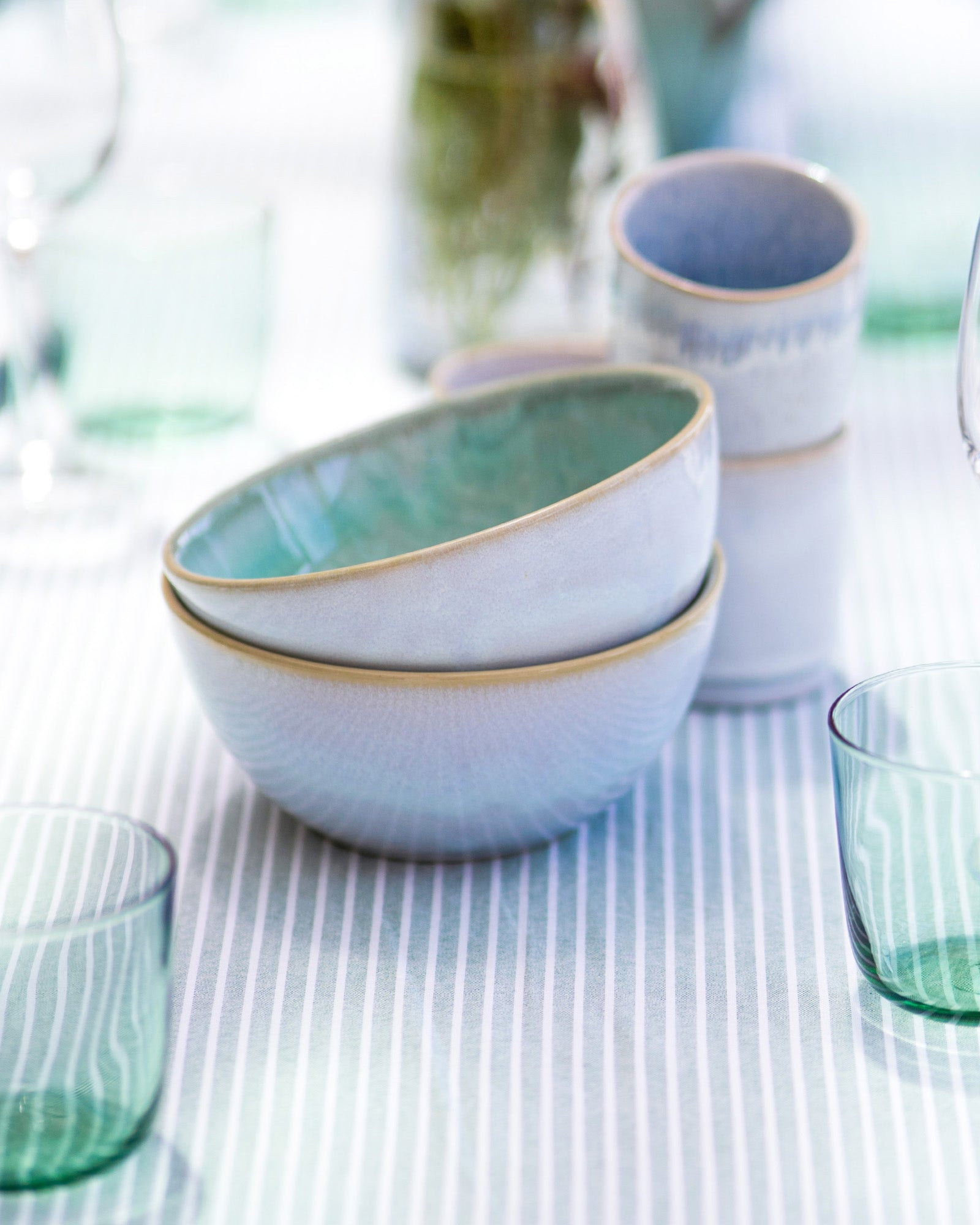 Stapel von Keramikschalen auf gestreifter Tischdecke, umgeben von grünen Gläsern und verschwommenem Hintergrund.