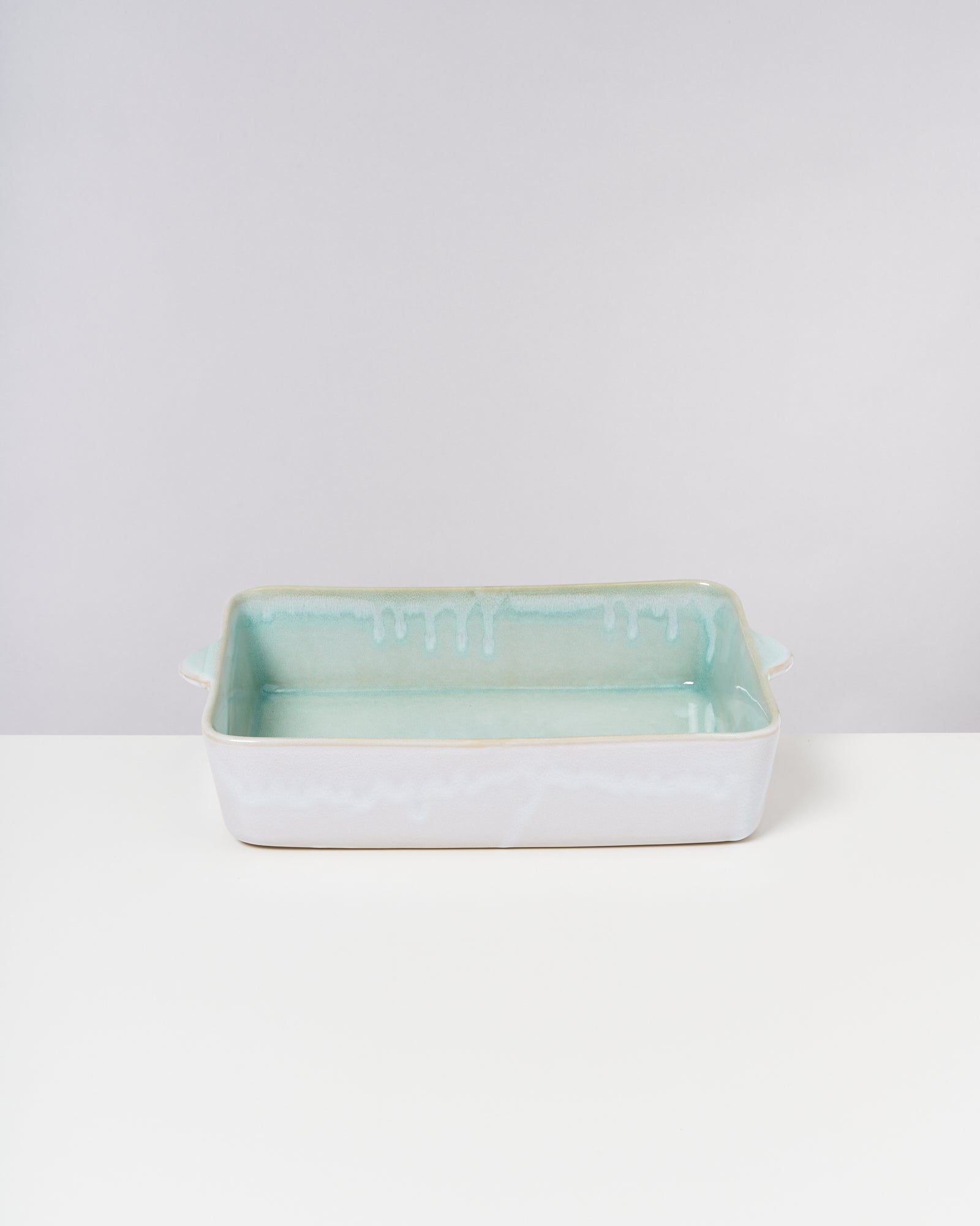 Rechteckige, blassgrüne Keramikbackform mit Henkeln steht auf einem weißen Tisch vor grauem Hintergrund.
