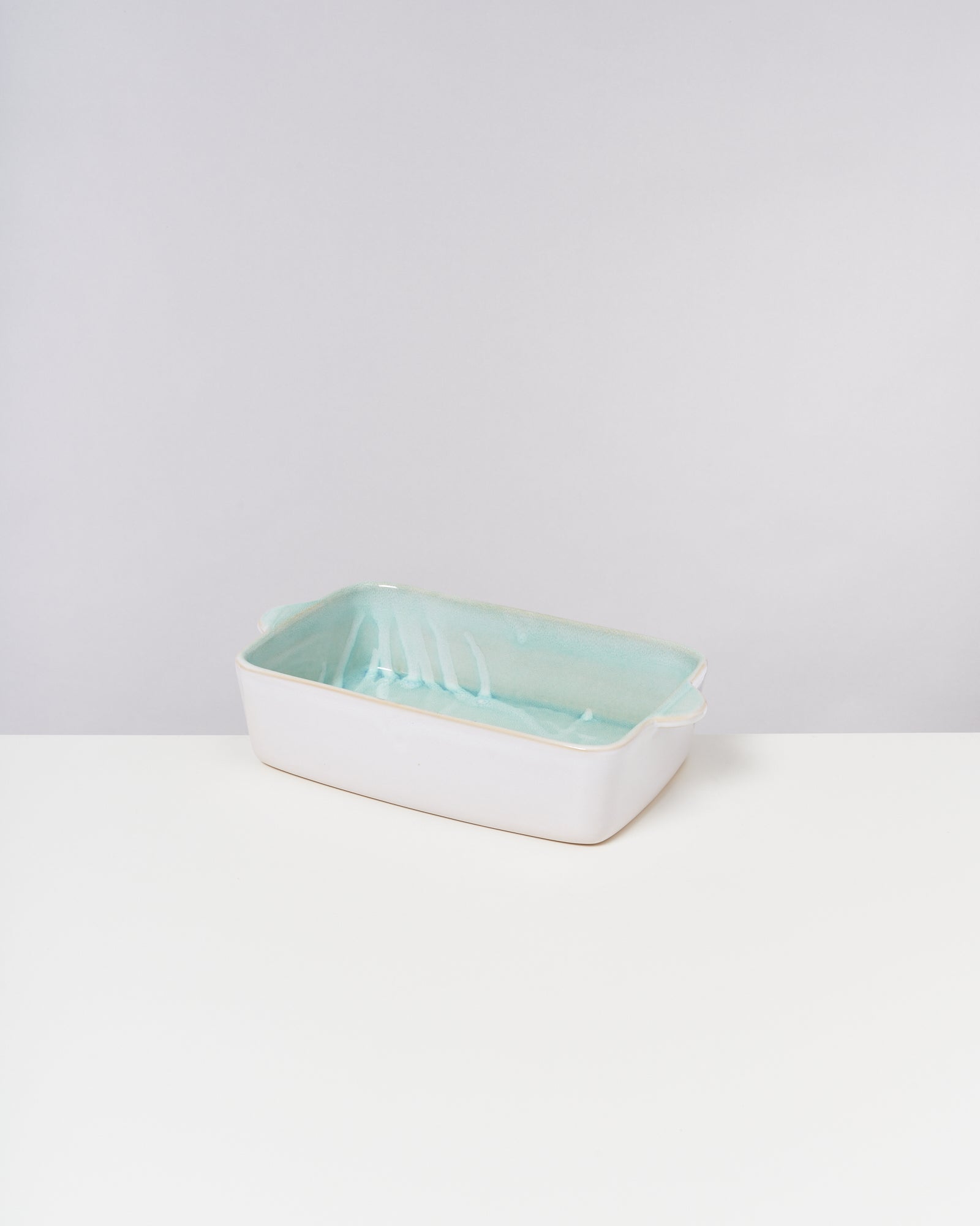 Weiße rechteckige Keramikschale mit türkisfarbenem Inneren und kleinen Griffen, steht auf weißer Oberfläche.