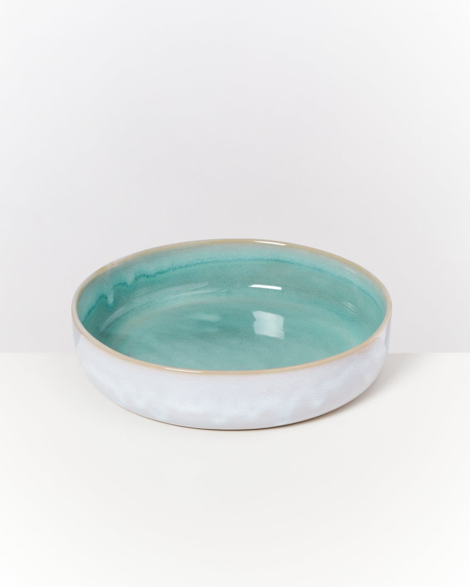 Flache, türkisfarbene Keramikschale mit einem weißen, leicht strukturierten Rand, auf einem weißen Hintergrund.