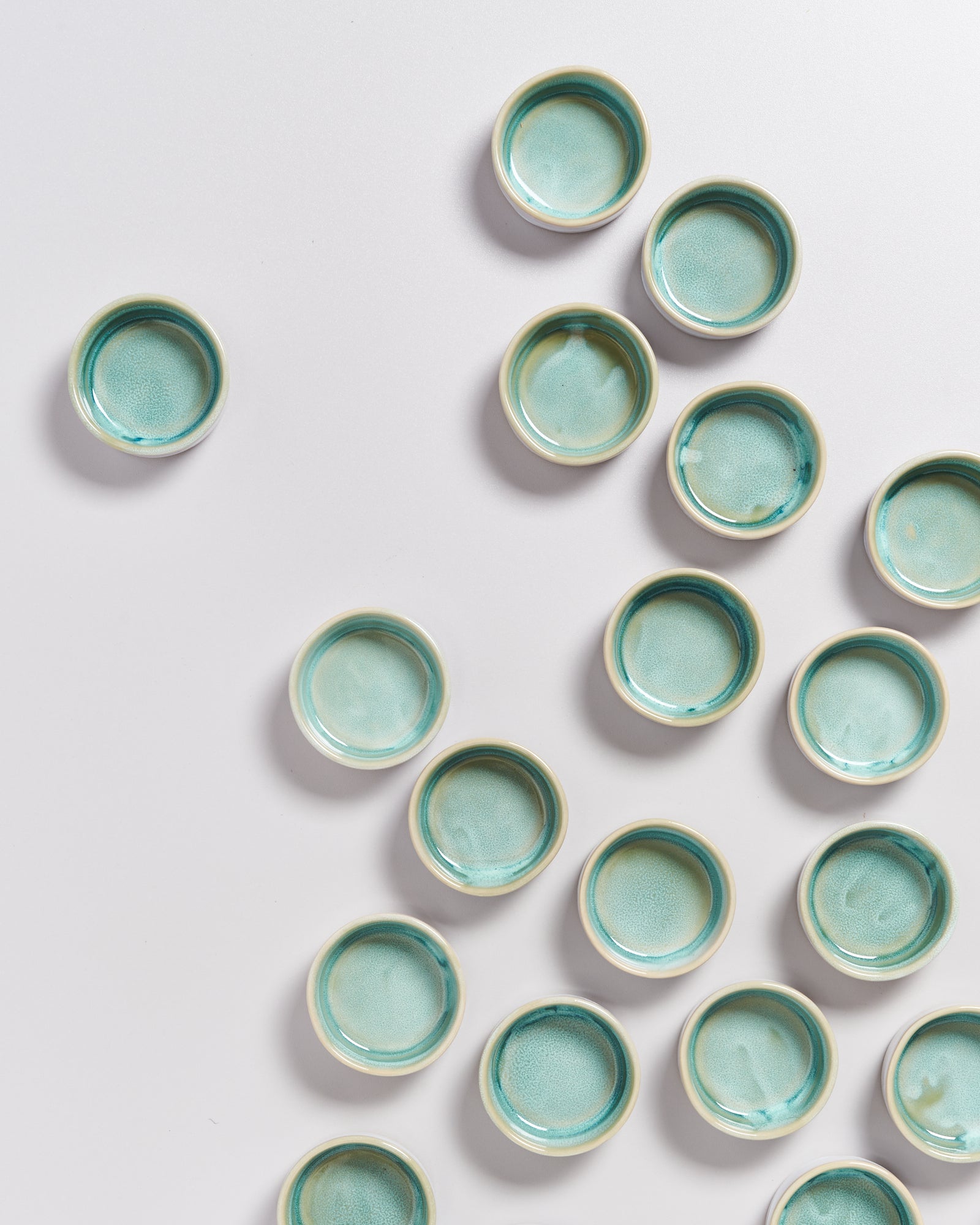 Mehrere grün-blaue, runde Keramikschalen sind auf einer hellen Oberfläche verstreut angeordnet.