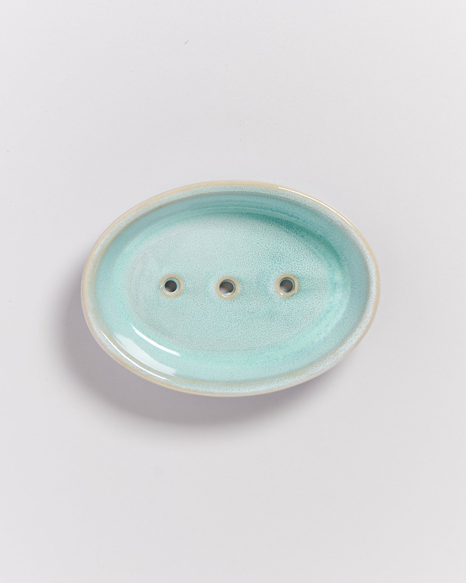 Ovale Seifenschale aus türkisfarbenem Keramik mit drei Löchern, auf einer weißen Oberfläche.