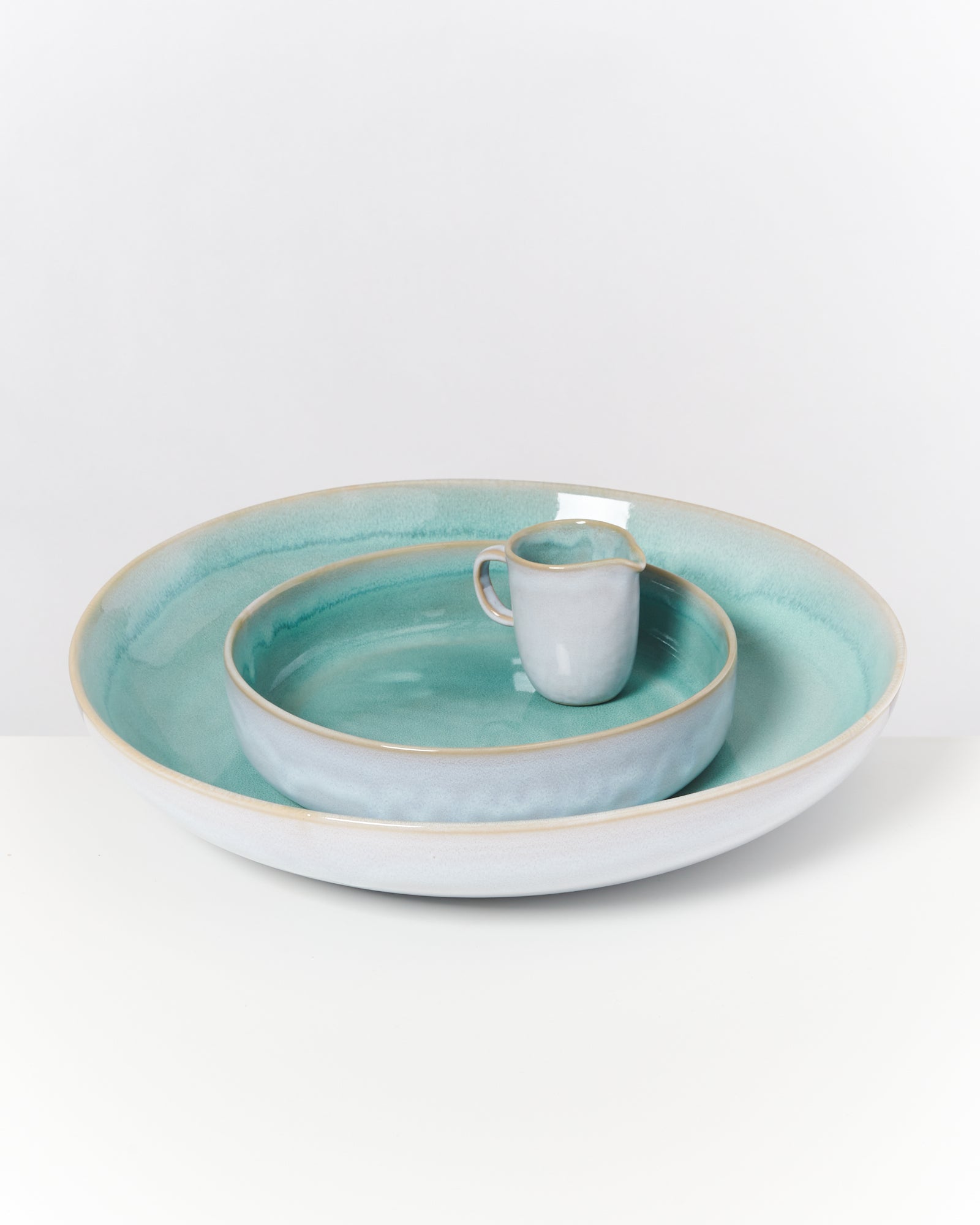 Drei Keramikteller in verschiedenen Größen und einem kleinen Kännchen, alle in Pastellgrün und Hellgrau auf weißem Hintergrund.