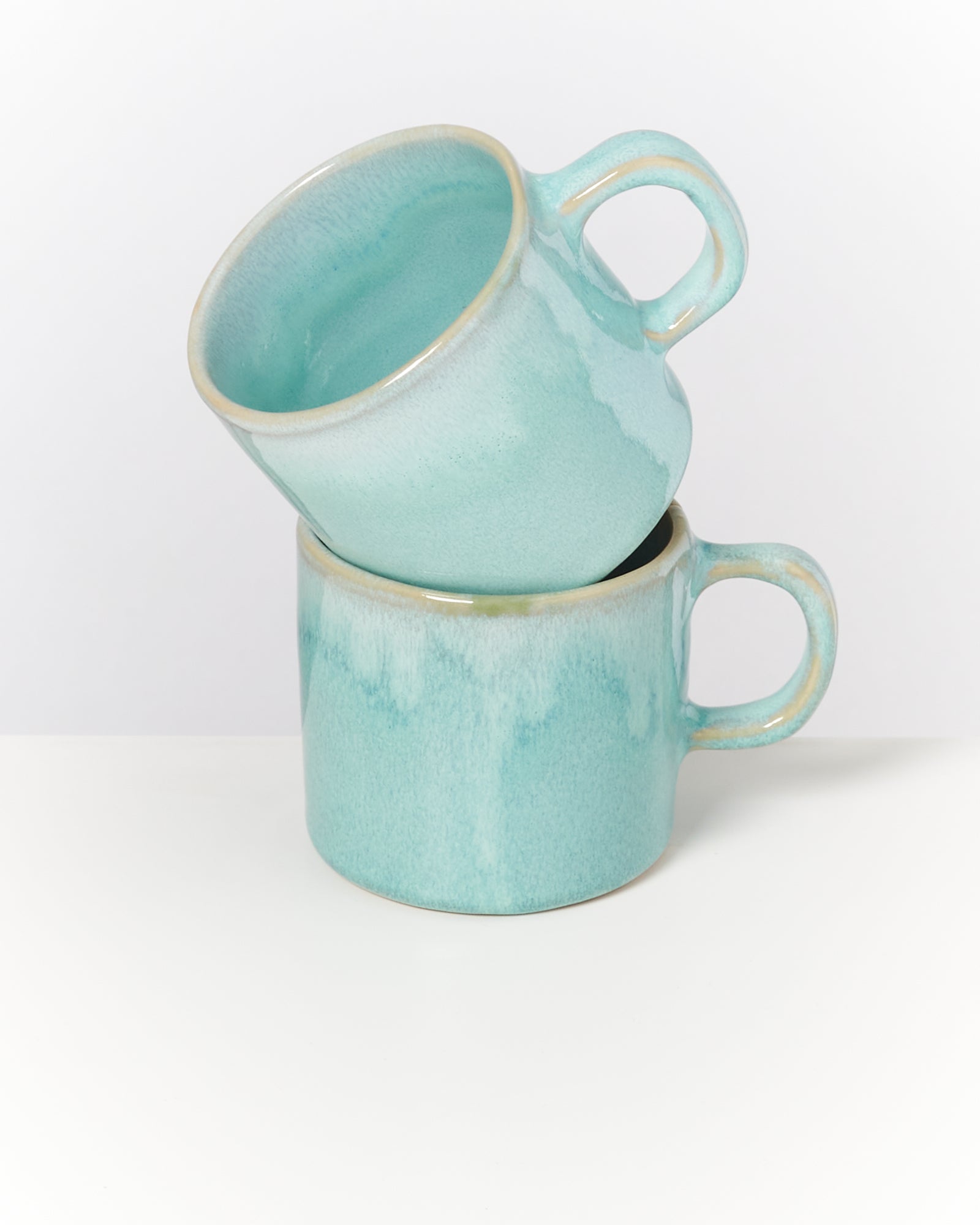 Zwei türkisfarbene Keramiktassen, eine liegt auf der anderen, auf weißem Hintergrund.