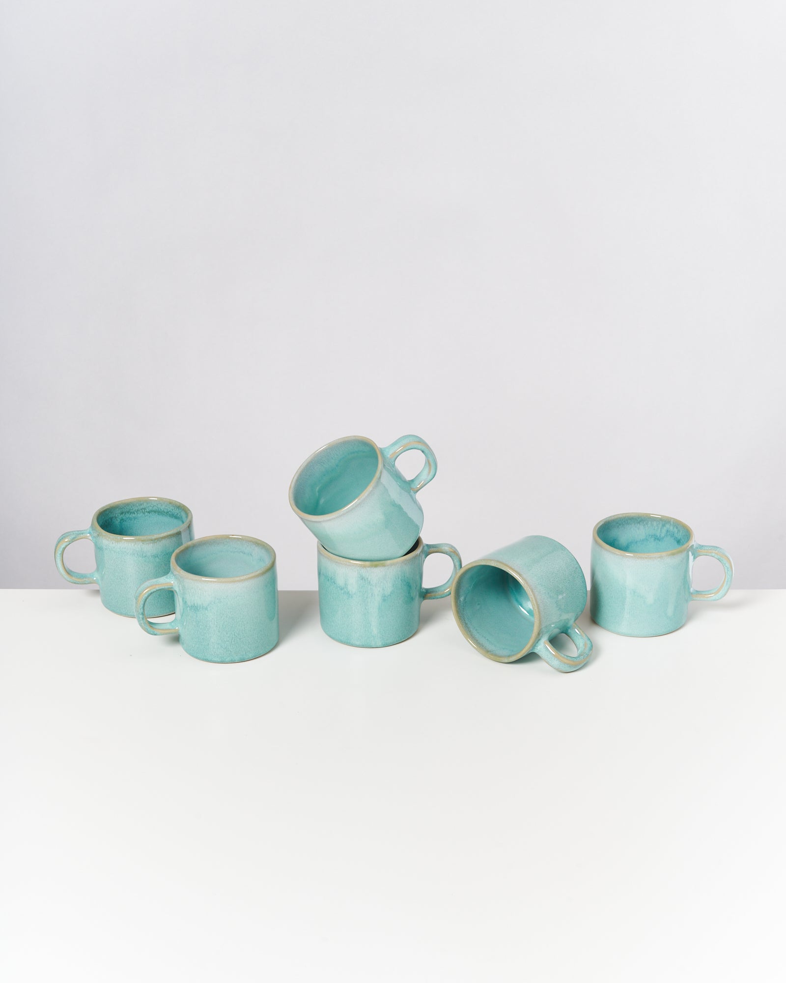 Sechs türkisfarbene Keramiktassen auf einem weißen Tisch, eine Tasse liegt seitlich.