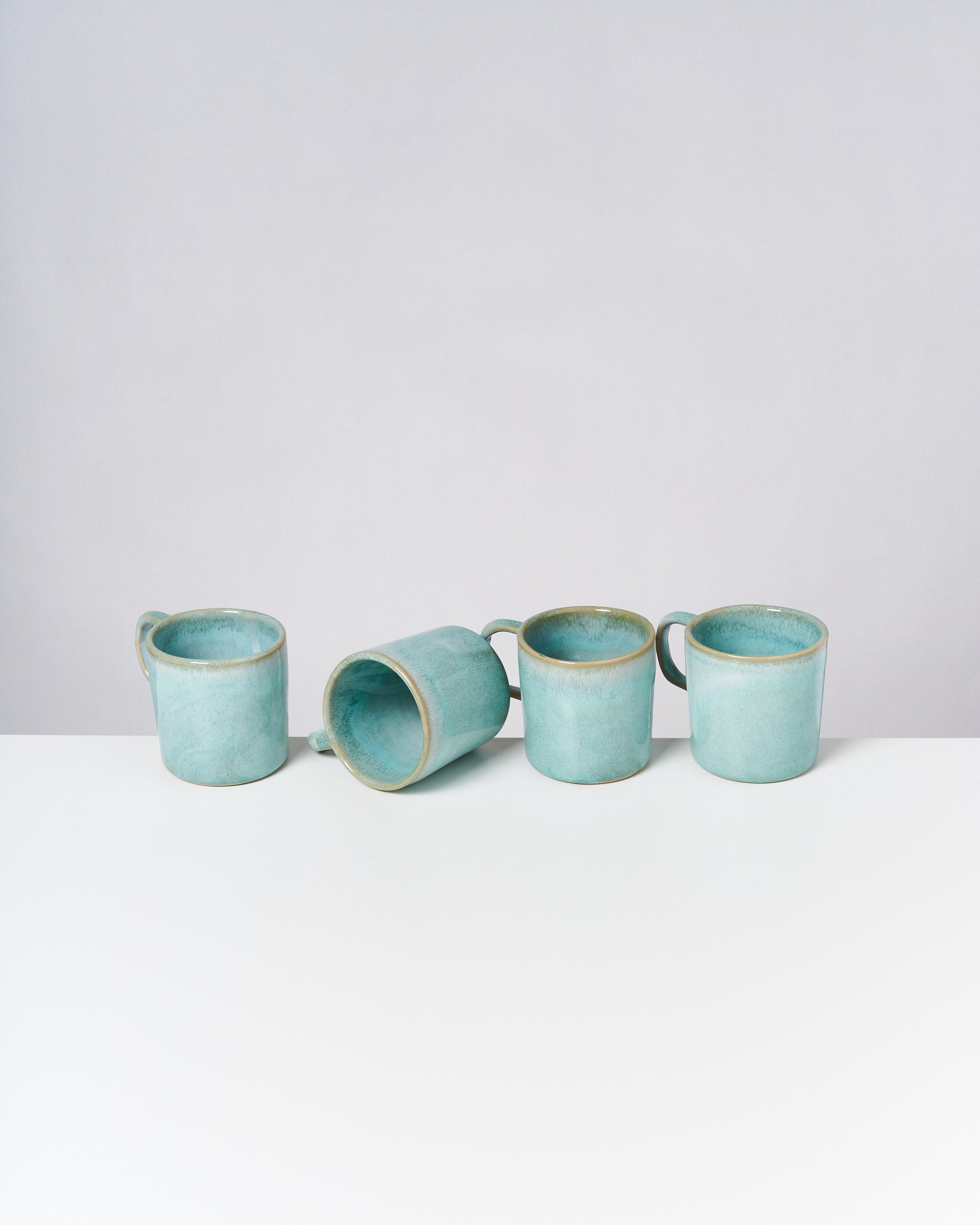 Vier türkisfarbene Tassen stehen auf einem weißen Tisch, eine davon liegt auf der Seite.