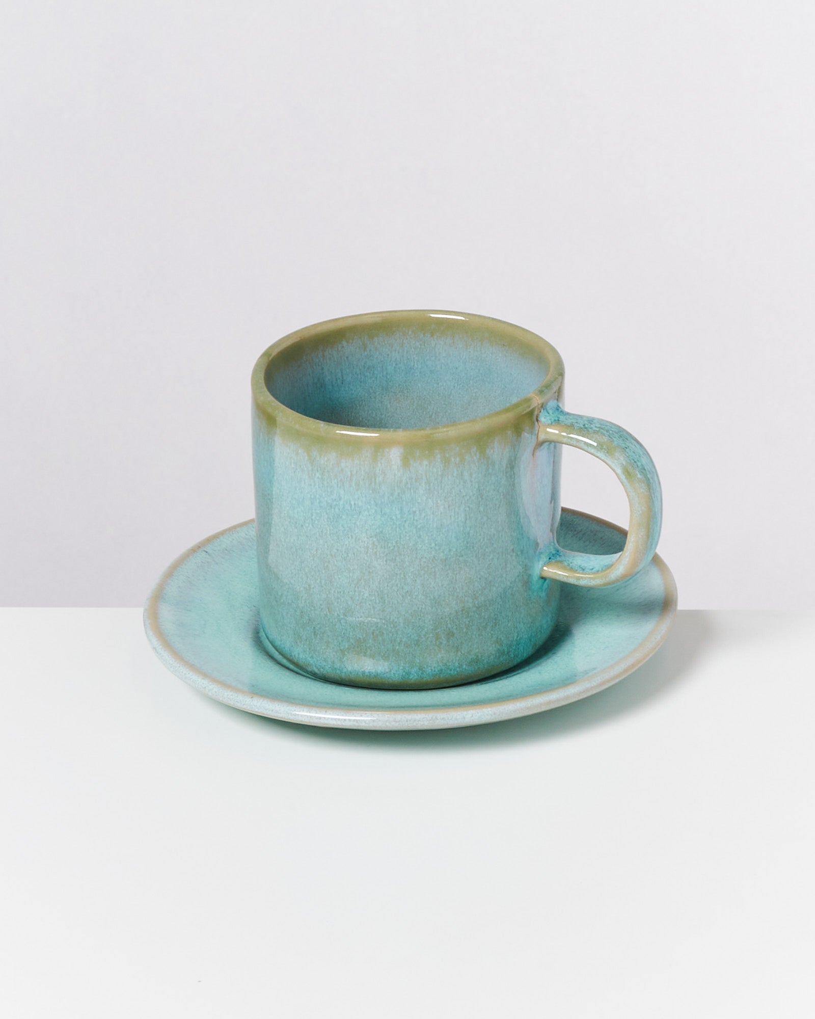 Türkisfarbene Keramiktasse auf passender Untertasse vor weißem Hintergrund.