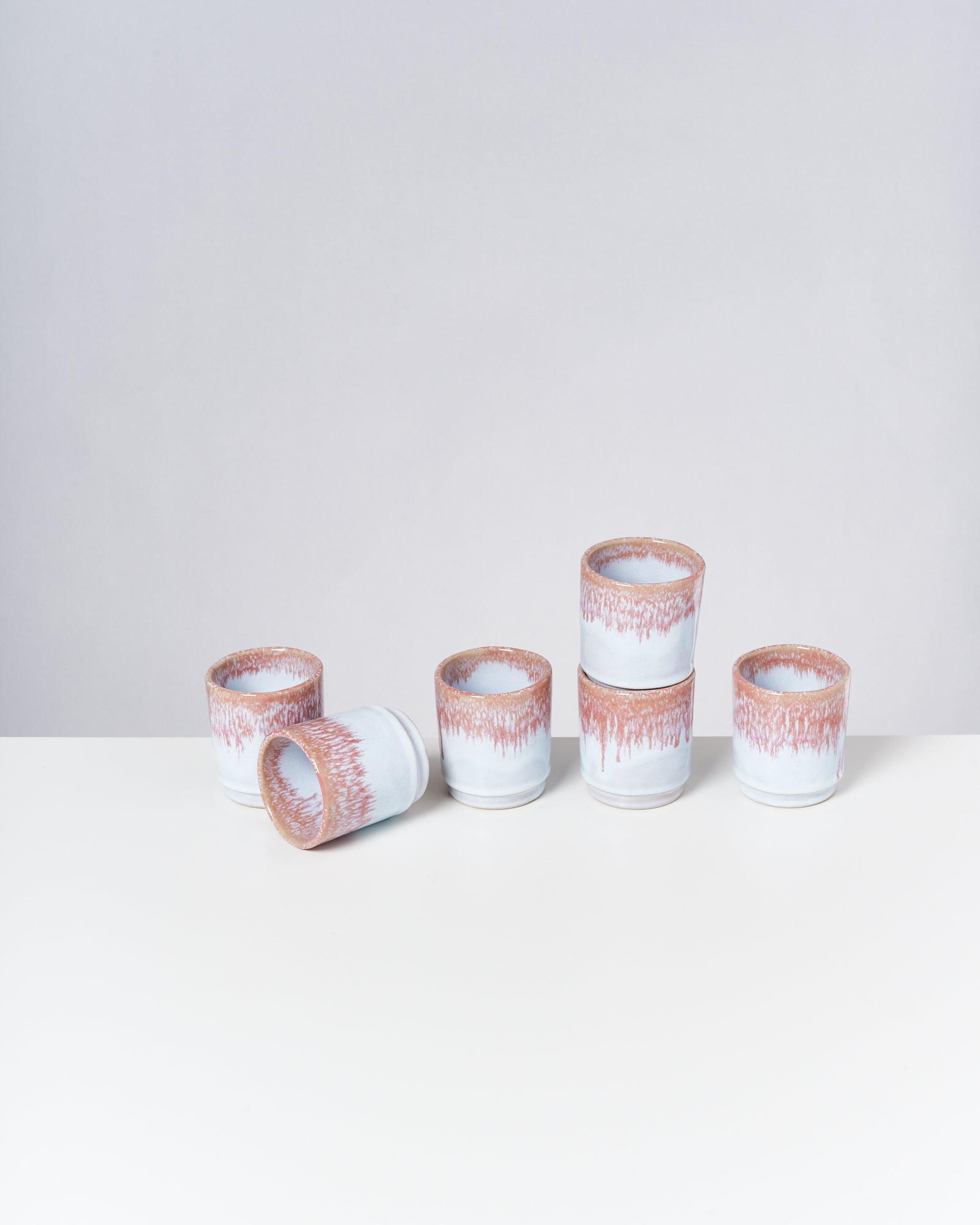 Fünf Keramiktassen mit weißen und roten Mustern stehen auf einem weißen Tisch, wobei eine Tasse liegt.