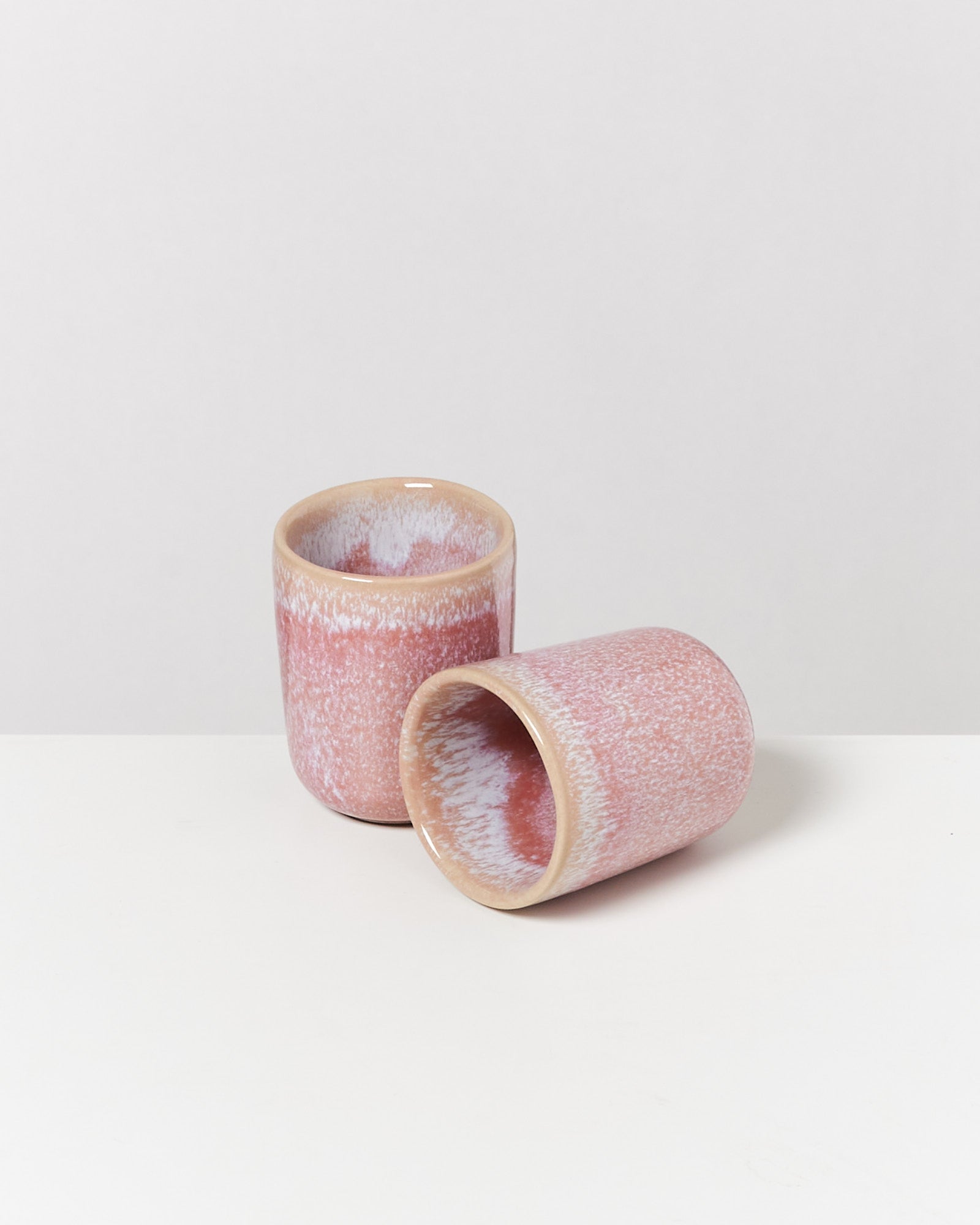 Zwei rosafarbene Keramikbecher auf einer weißen Fläche, einer steht, der andere liegt.