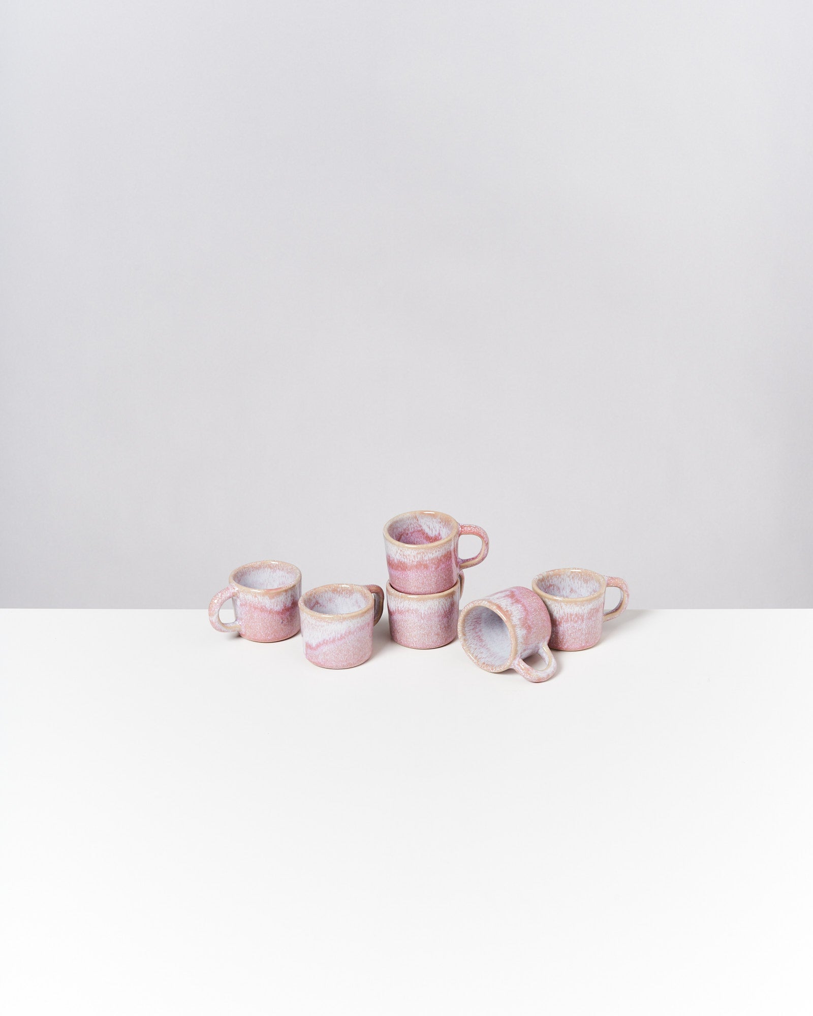 Sechs rosafarbene Kaffeetassen aus Keramik stehen auf einem weißen Tisch vor grauem Hintergrund.