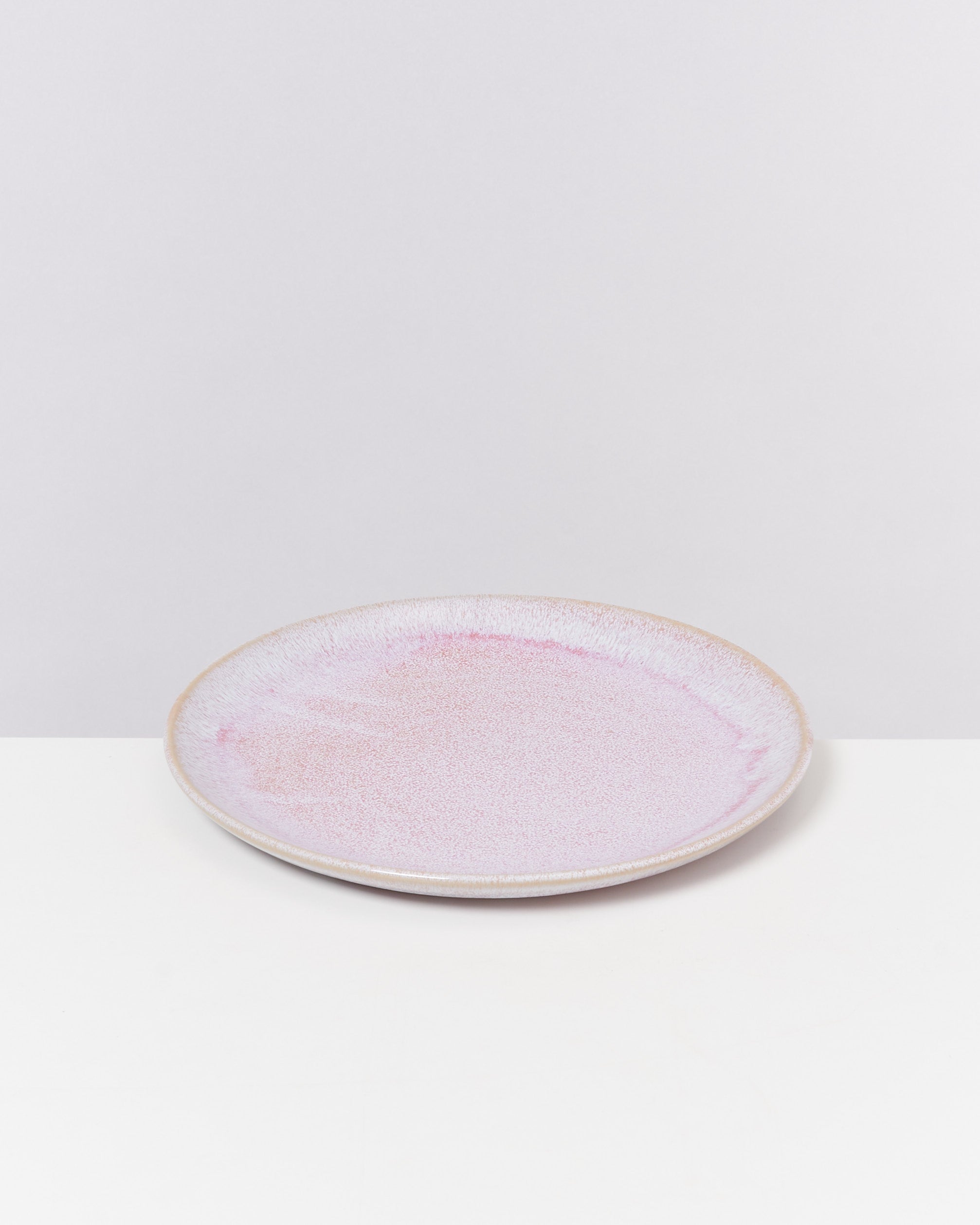 Runder, flacher Teller mit rosa-weißer Glasur, steht auf einer weißen Oberfläche vor grauem Hintergrund.