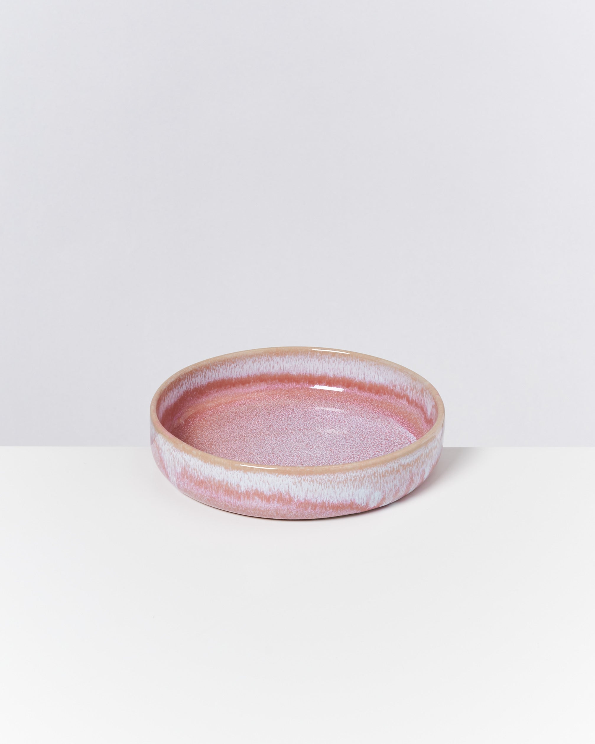 Runde, flache, rosa Keramikschale mit weißem Rand auf weißem Hintergrund.
