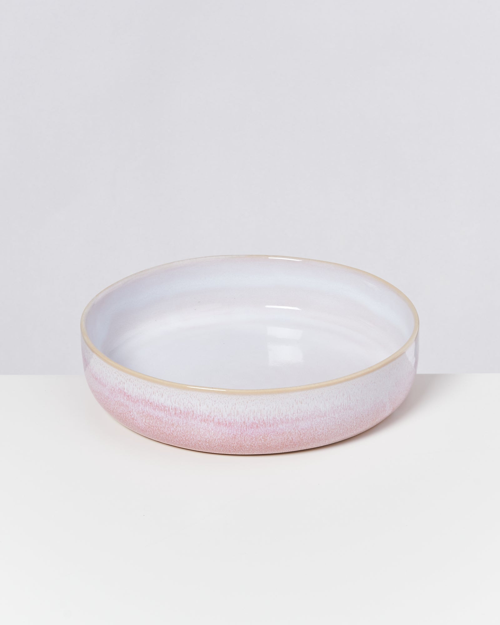 Flache, runde Schale aus Keramik in Pastellrosa und Weiß auf weißem Hintergrund.