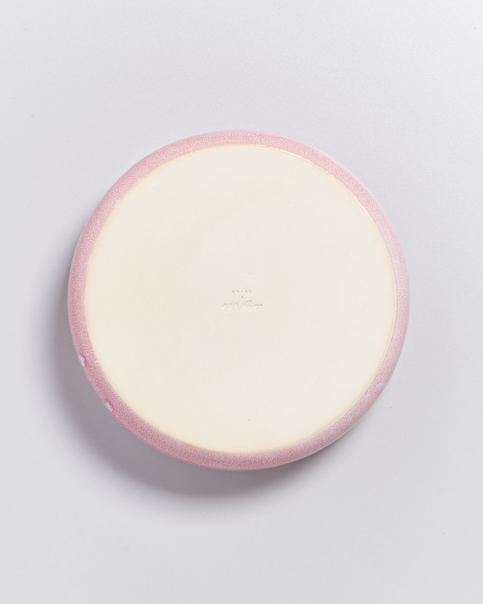 Runde, beige Unterseite eines Keramikobjekts mit rosa Rand vor grauem Hintergrund.