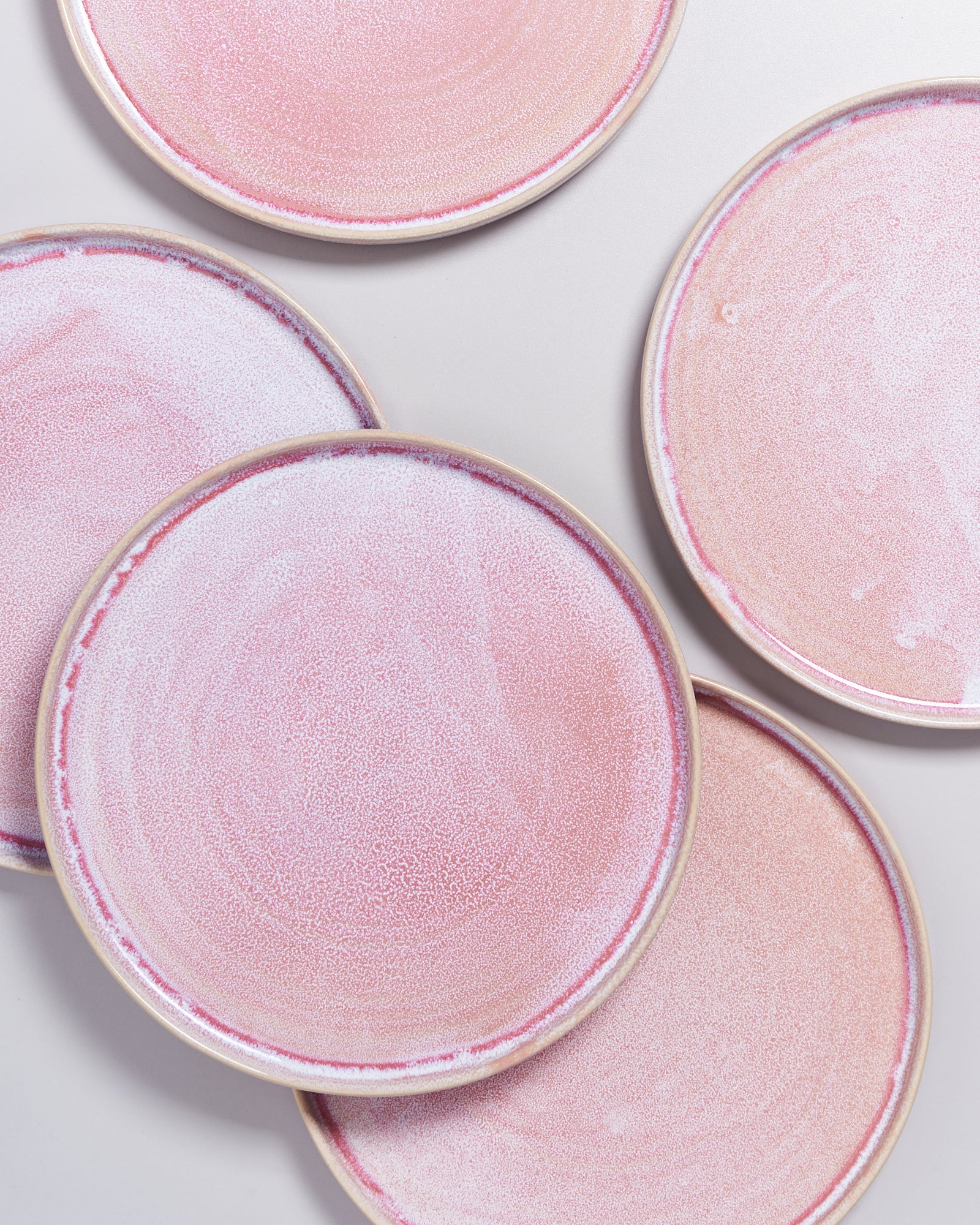 Fünf runde, rosafarbene Keramikteller mit einem gemusterten Design liegen unregelmäßig angeordnet auf einer weißen Oberfläche.