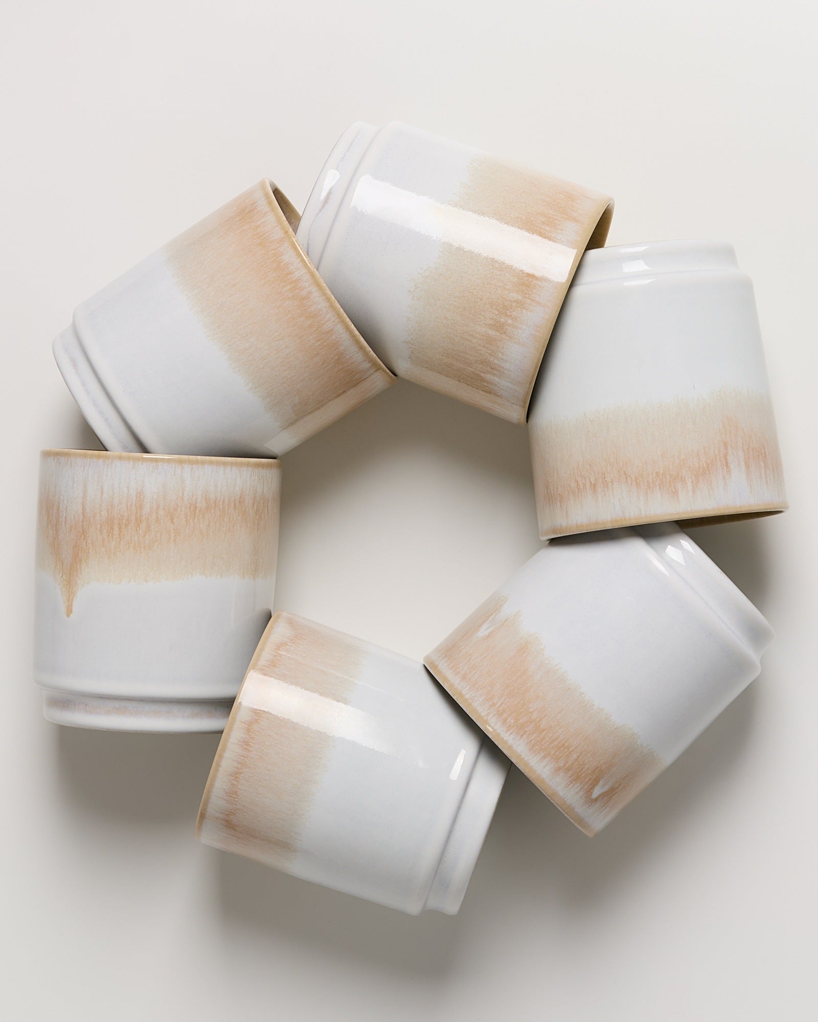 Sechs weiße Keramiktassen mit braunem Farbverlauf liegen ringförmig auf einem weißen Hintergrund.