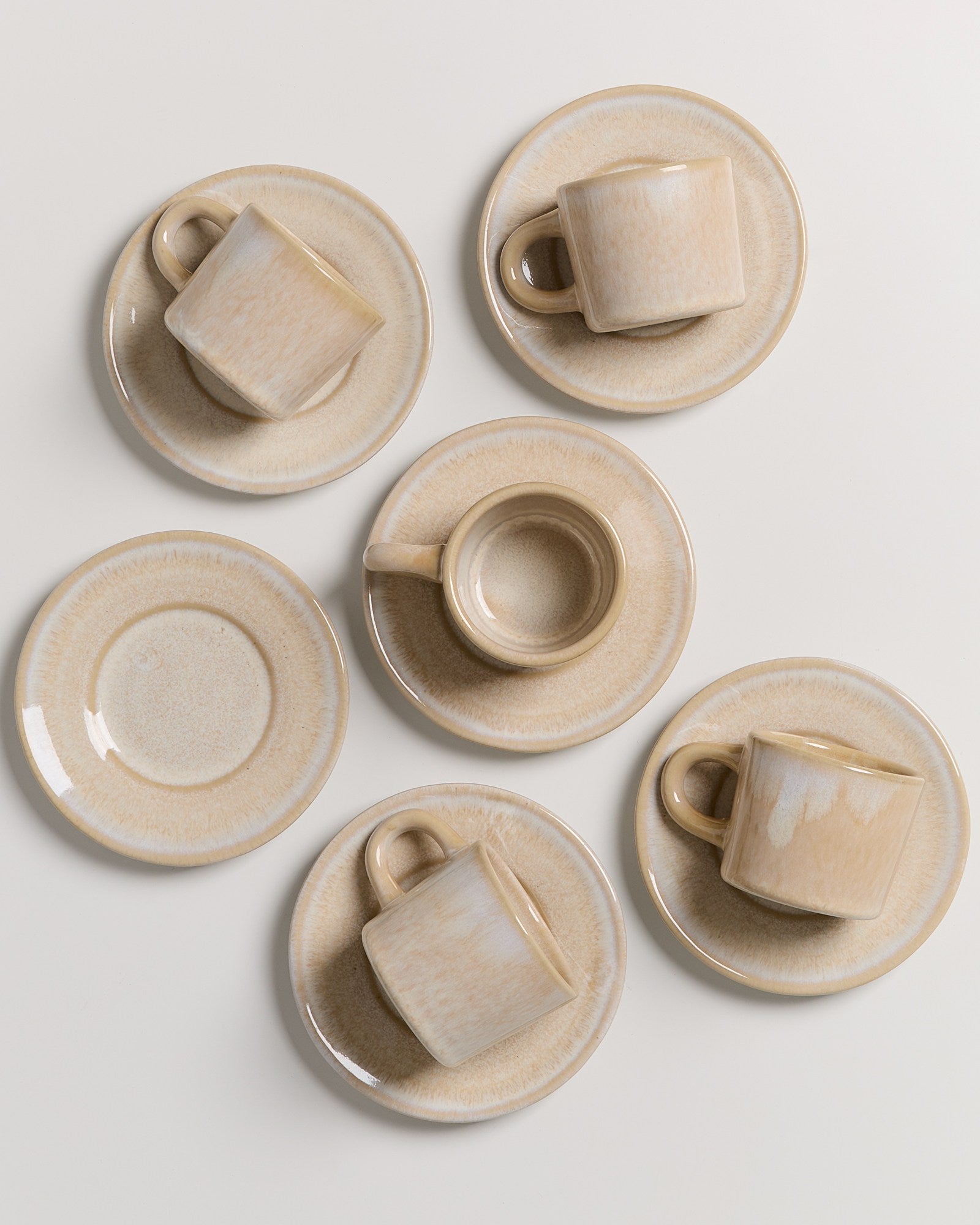 Sechs beigefarbene Kaffeetassen mit passenden Untertassen, symmetrisch auf einer weißen Fläche angeordnet.