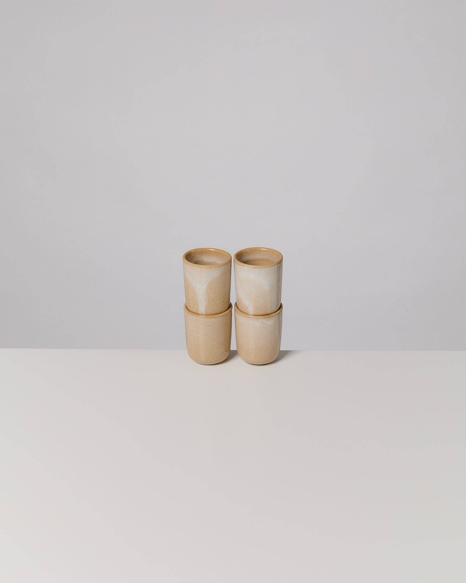 Vier stapelbare, beige Tassen aus Keramik stehen auf einer weißen Oberfläche vor einem grauen Hintergrund.