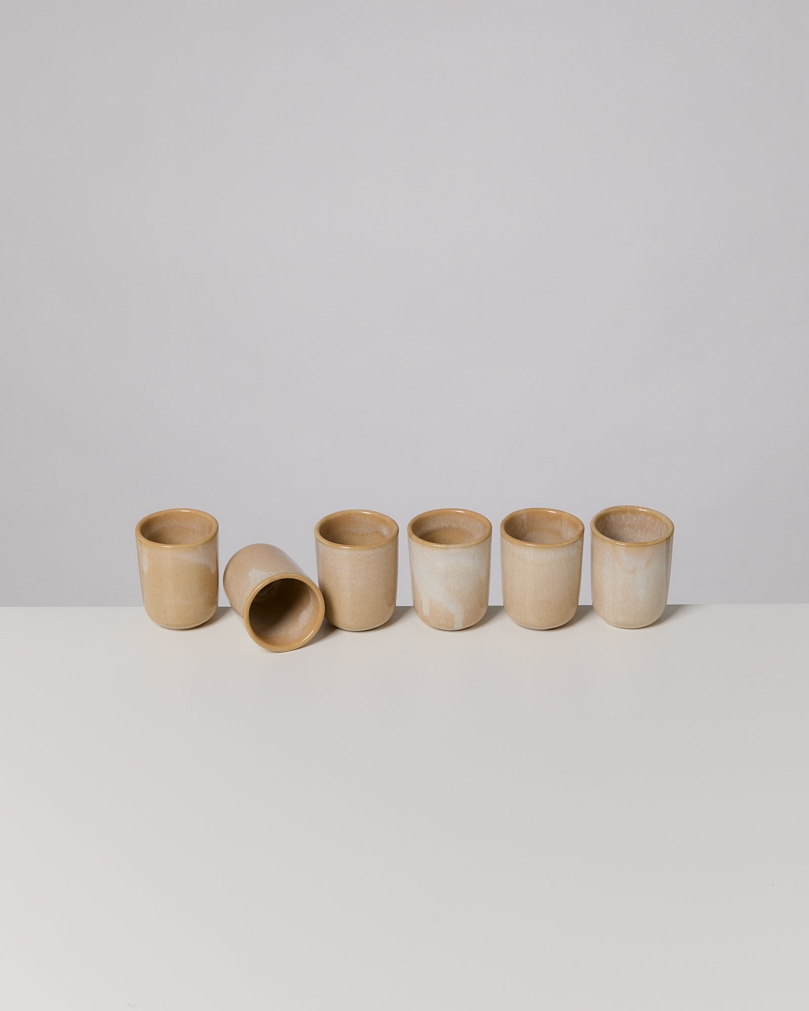 Fünf beige Keramiktassen stehen nebeneinander, eine ist umgekippt, auf einem weißen Tisch vor grauem Hintergrund.
