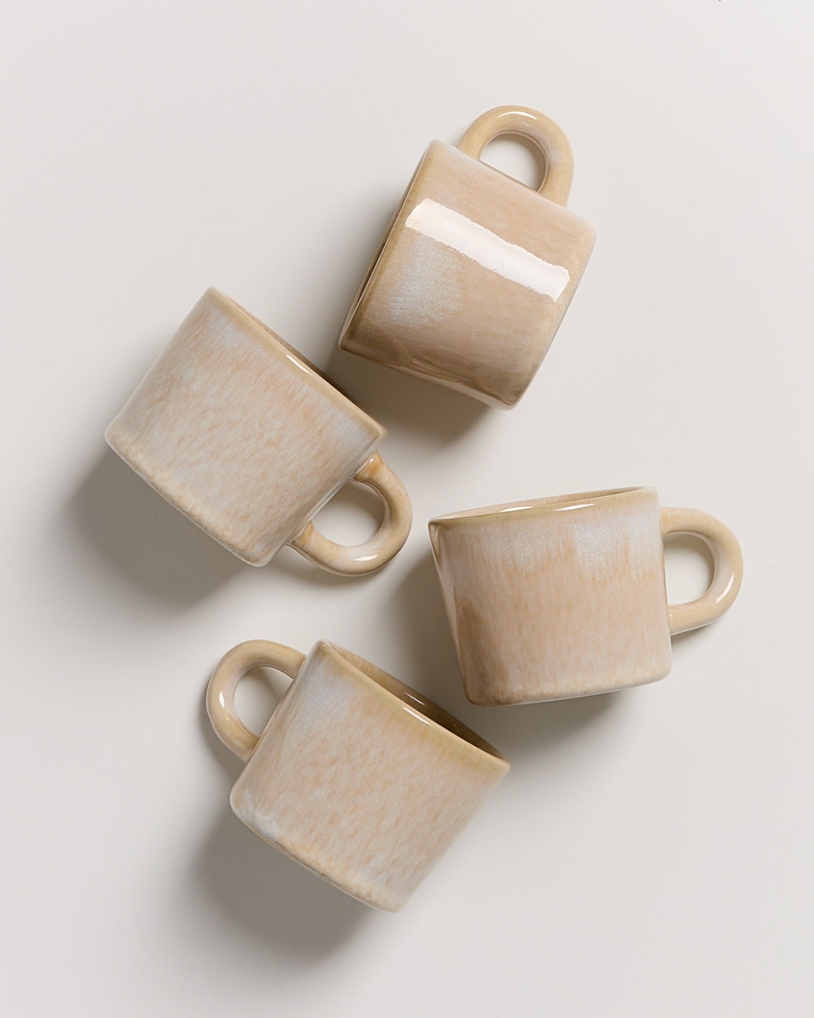 Vier beige Kaffeetassen mit Henkeln liegen auf einem hellen Untergrund.