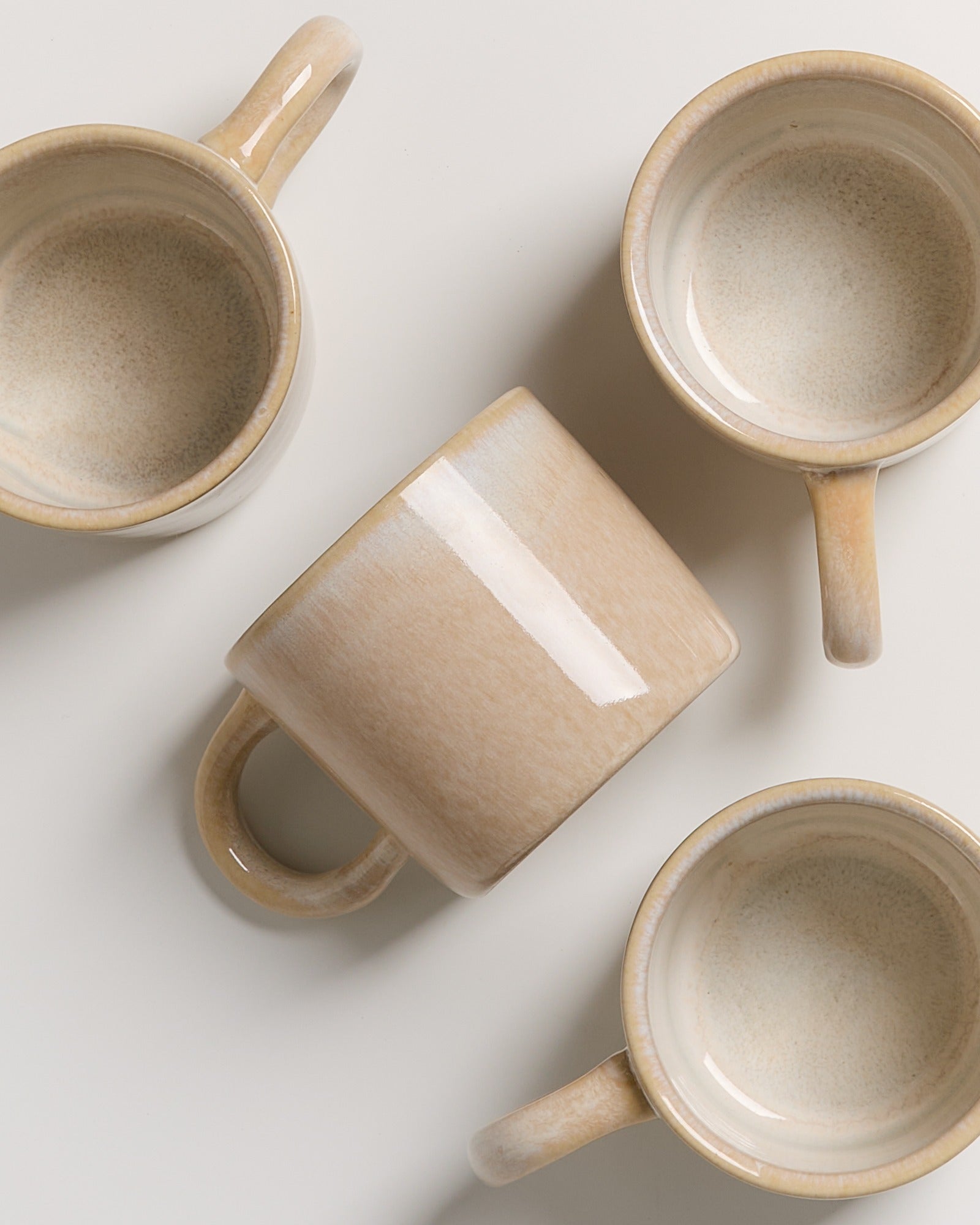 Vier beige Keramiktassen liegen auf einer hellen Oberfläche verteilt.