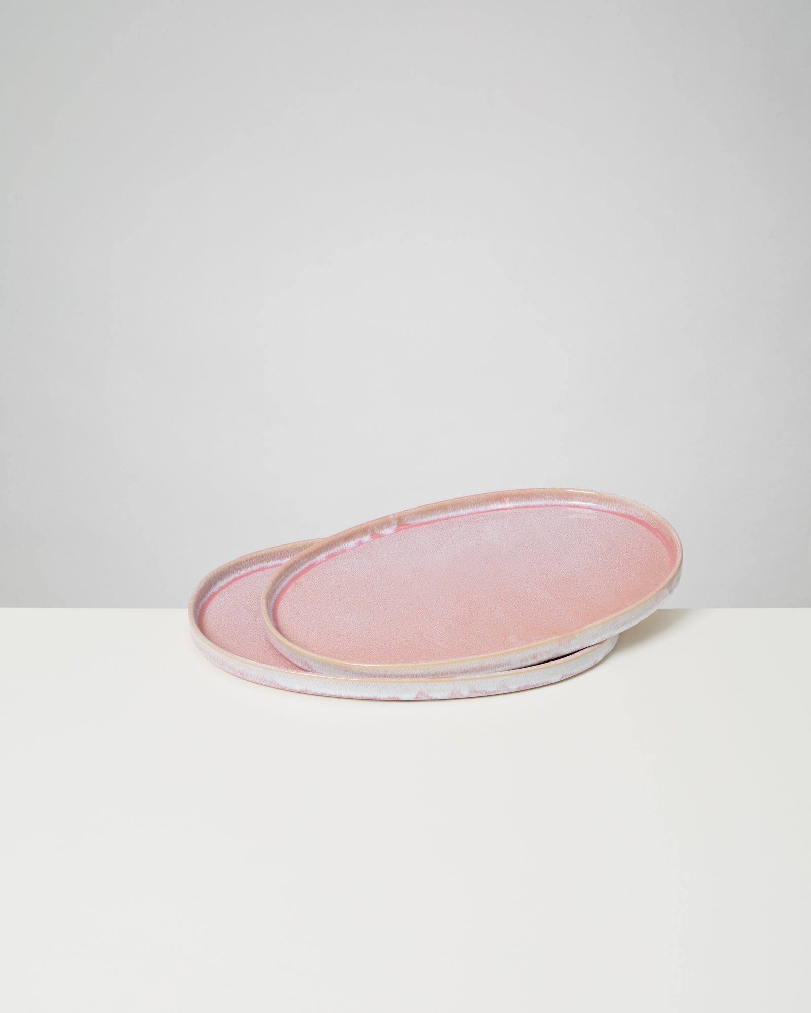 Zwei rosafarbene, ovale Keramikteller liegen übereinander auf einer weißen Fläche vor einem grauen Hintergrund.