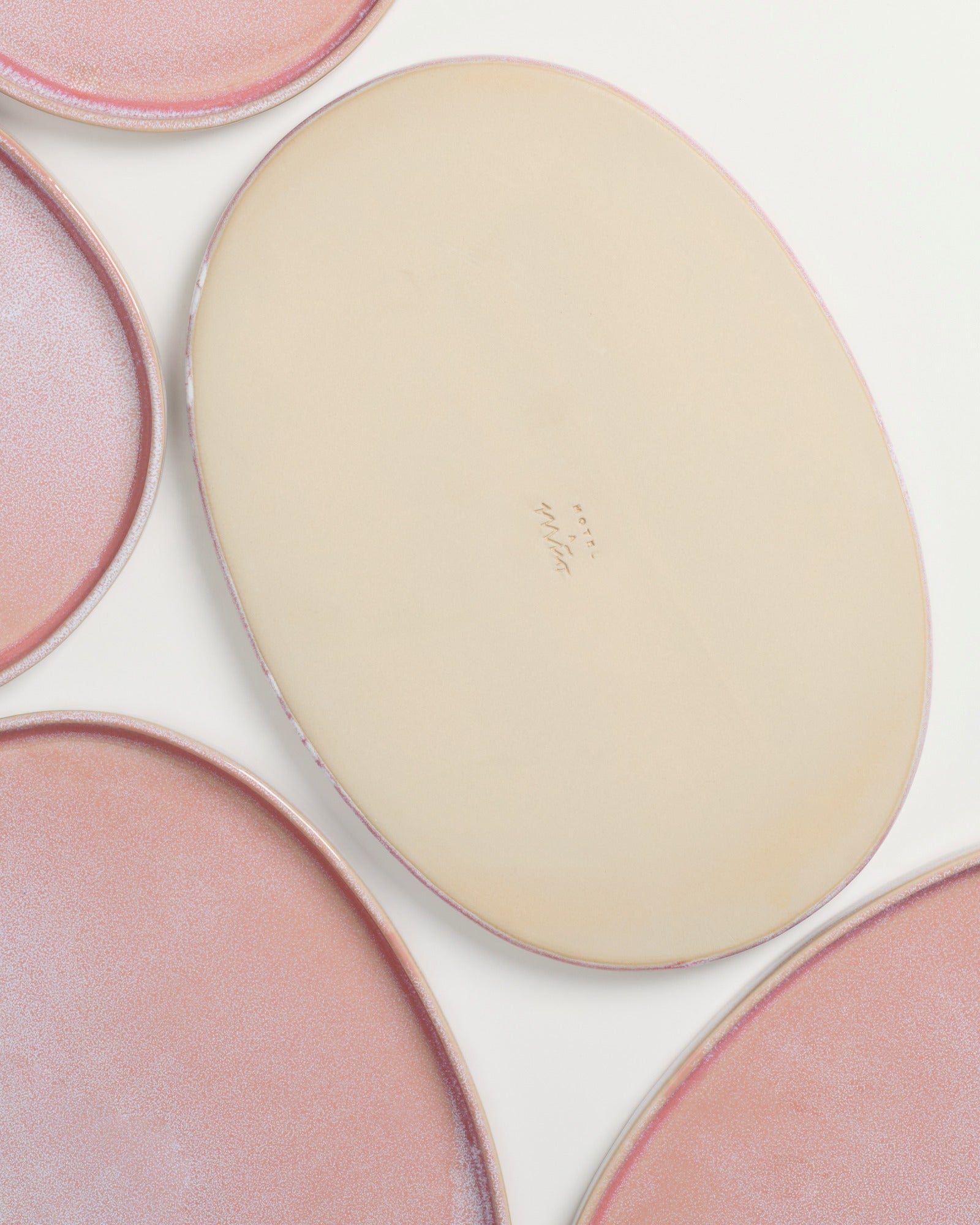 Ovale Teller mit rosafarbener Glasur auf einem hellen Hintergrund, ein Teller zeigt die unglasierte Rückseite mit Text.