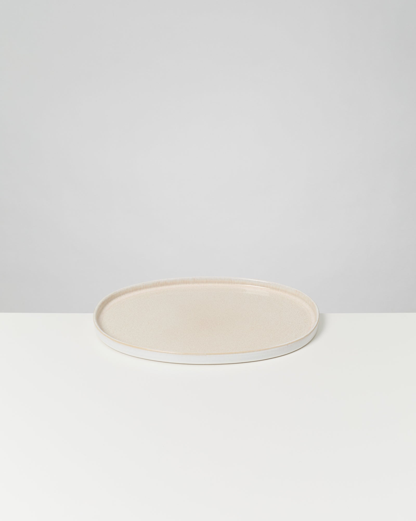 Ein flacher, runder, cremefarbener Teller steht auf einer weißen Oberfläche vor grauem Hintergrund.