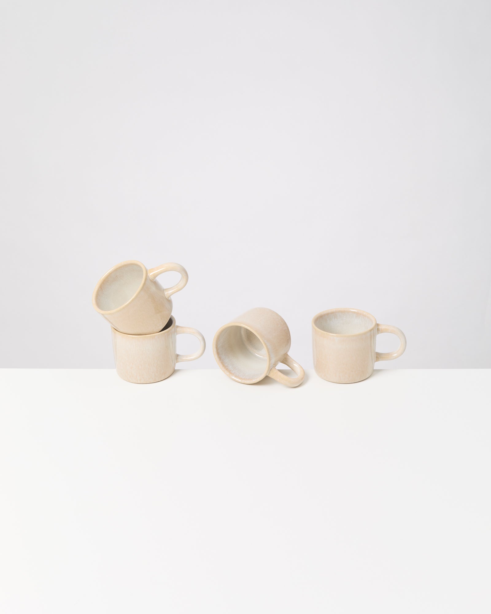 Vier beige Tassen, drei aufrecht, eine seitlich liegend, auf einem weißen Tisch vor einem weißen Hintergrund.