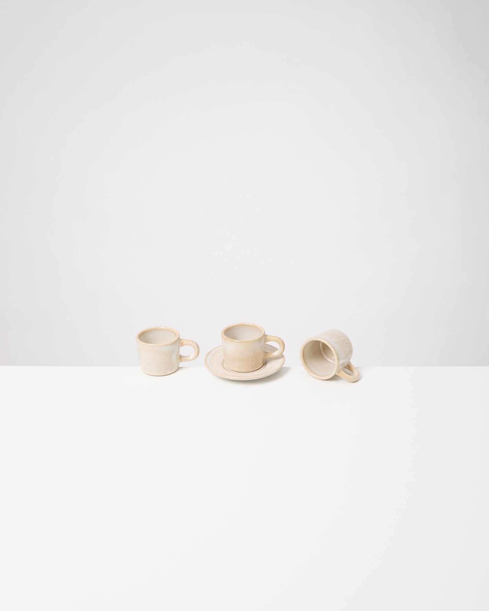Drei beigefarbene Kaffeetassen stehen auf einem weißen Tisch, eine auf einem Unterteller, eine liegt umgedreht.