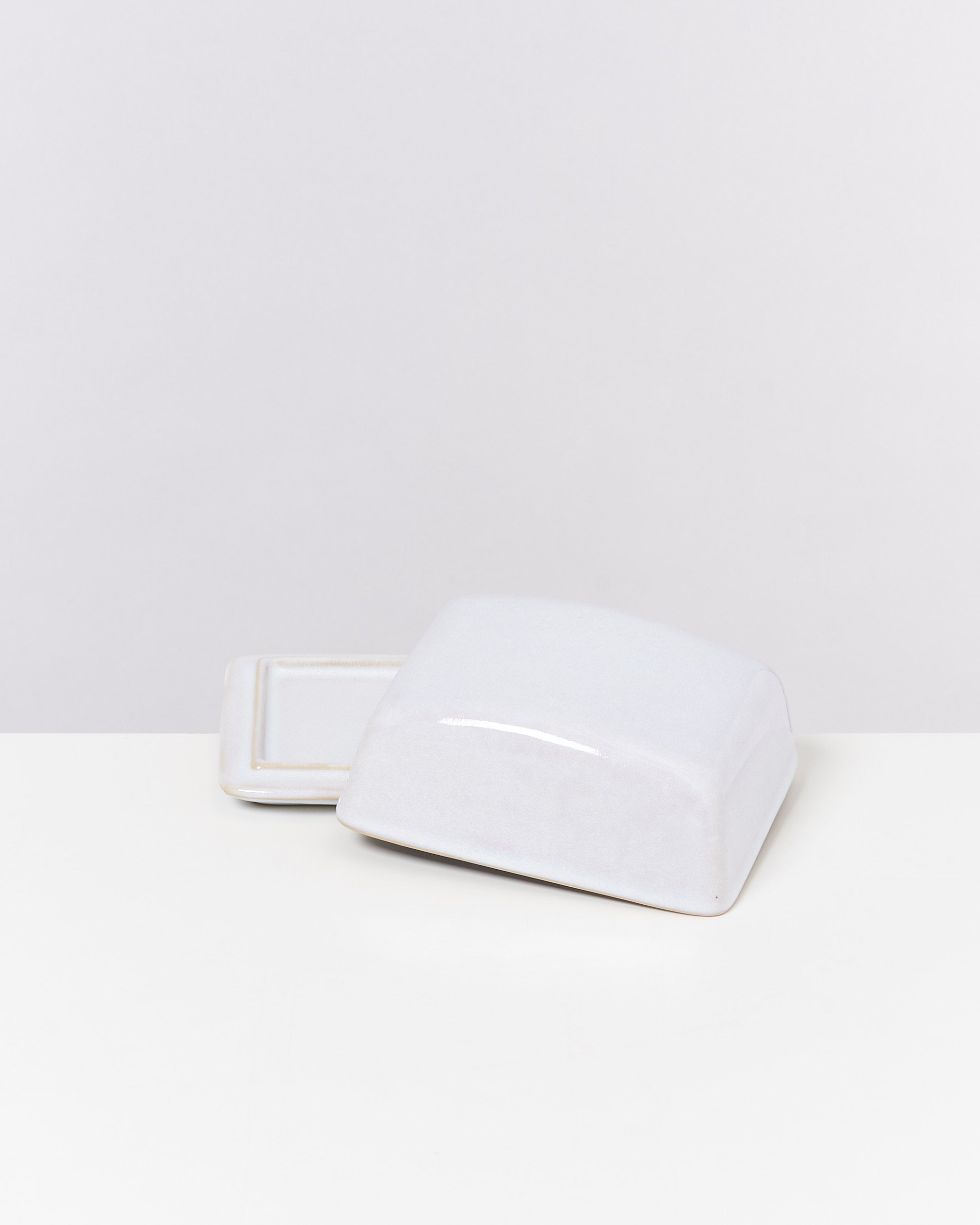 Weiße Butterdose aus Keramik mit abgenommenem Deckel auf einem weißen Tisch vor einem hellgrauen Hintergrund.