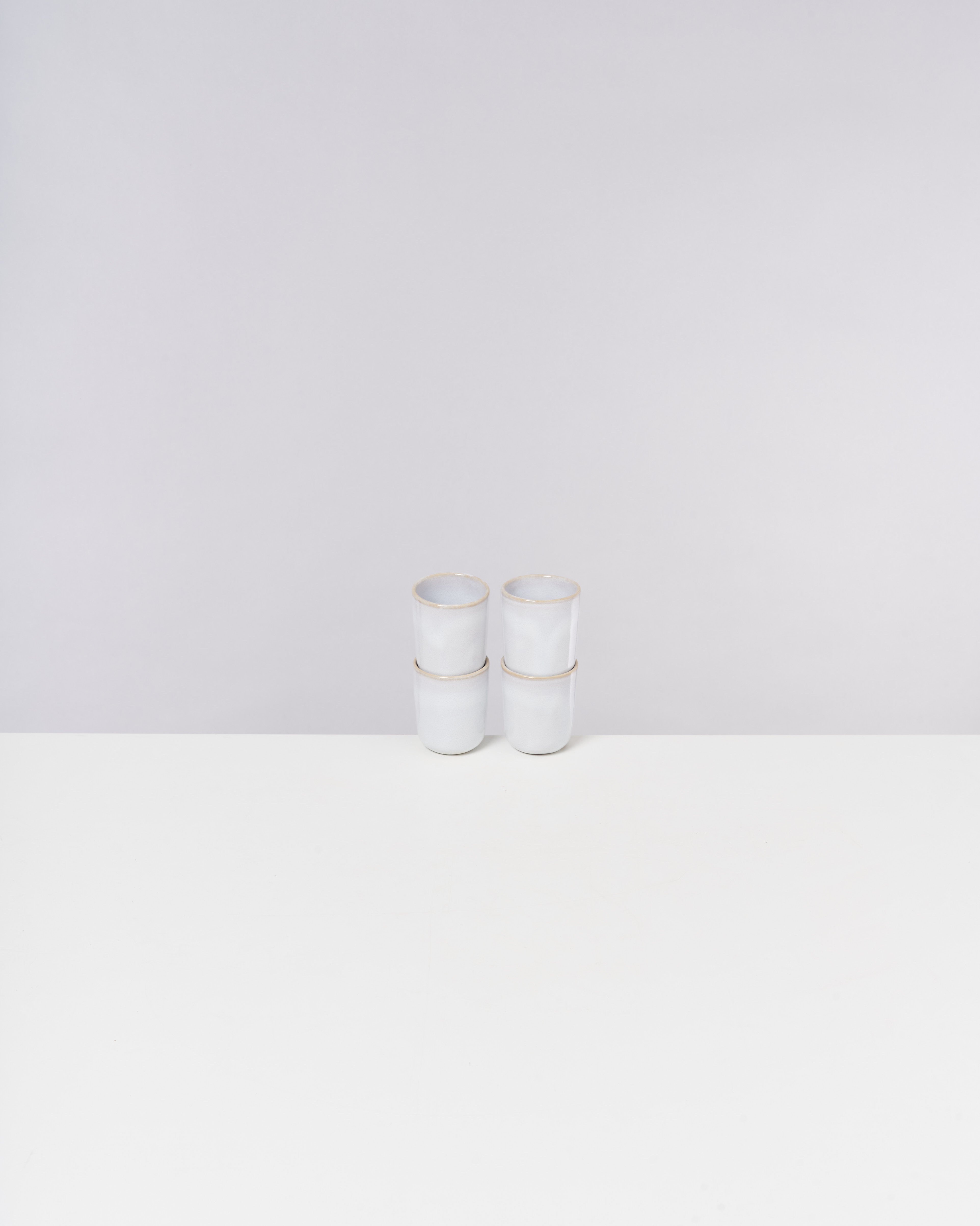 Vier gestapelte weiße Becher mit goldenen Rändern auf einem weißen Tisch vor grauem Hintergrund.
