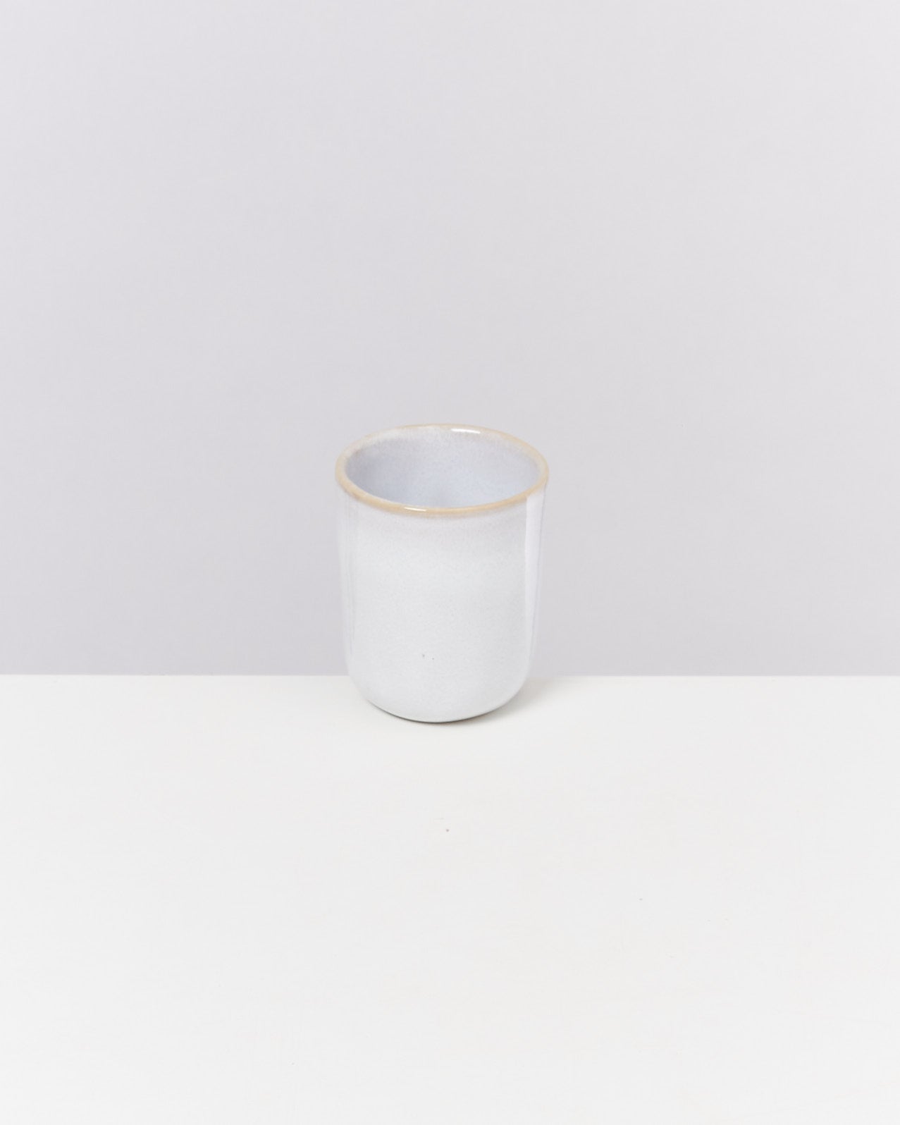 Ein weißer Keramikbecher mit leichtem Glanz steht auf einem weißen Tisch vor einem grauen Hintergrund.