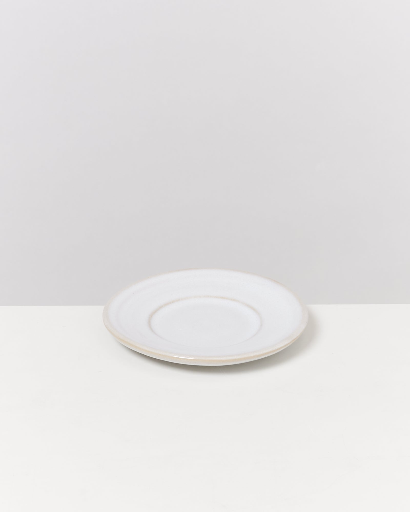 Ein weißer, flacher Teller steht auf einer weißen Oberfläche vor einem hellgrauen Hintergrund.