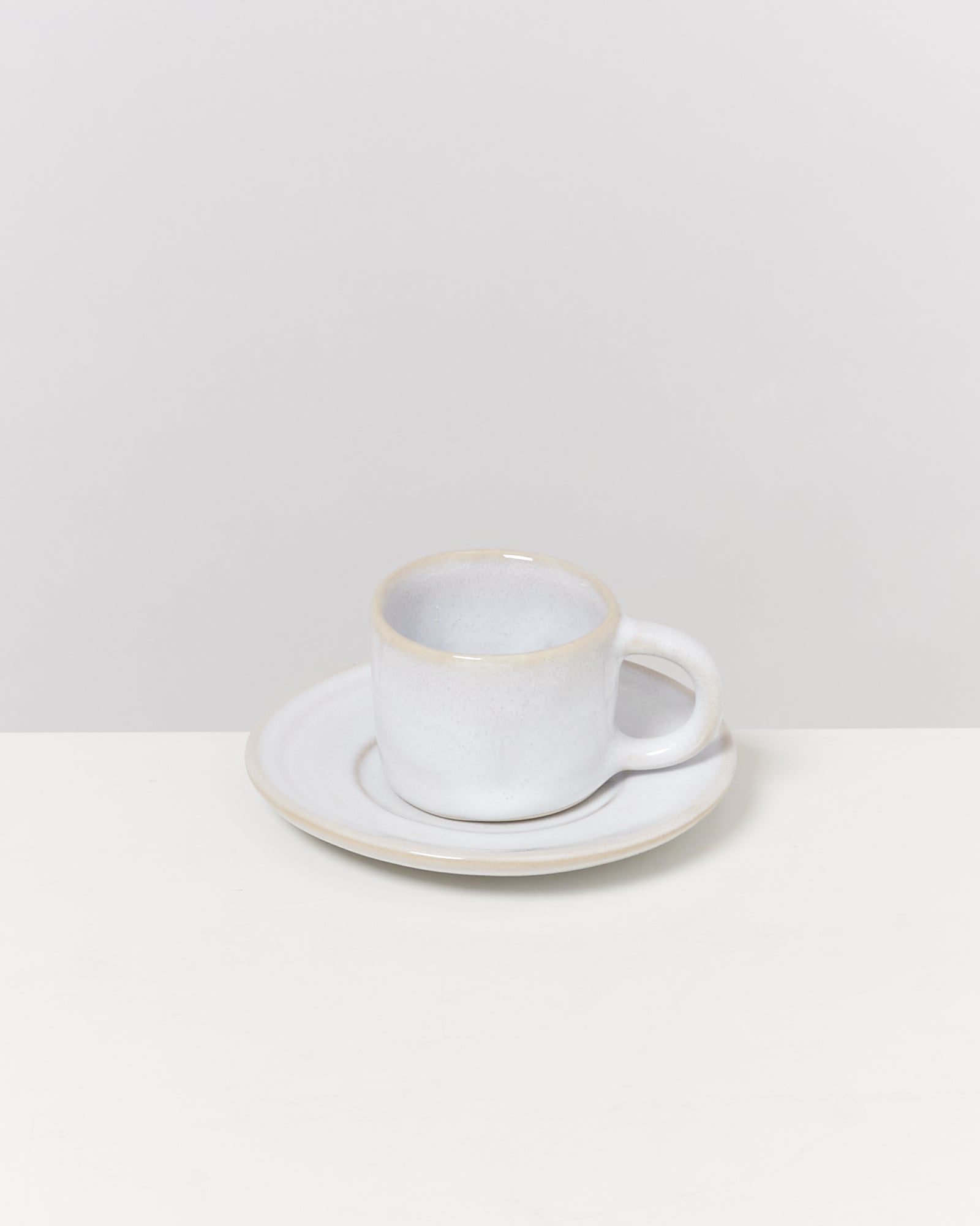 Weiße Keramiktasse mit Untertasse auf einem hellen Tisch vor einem neutralen Hintergrund.