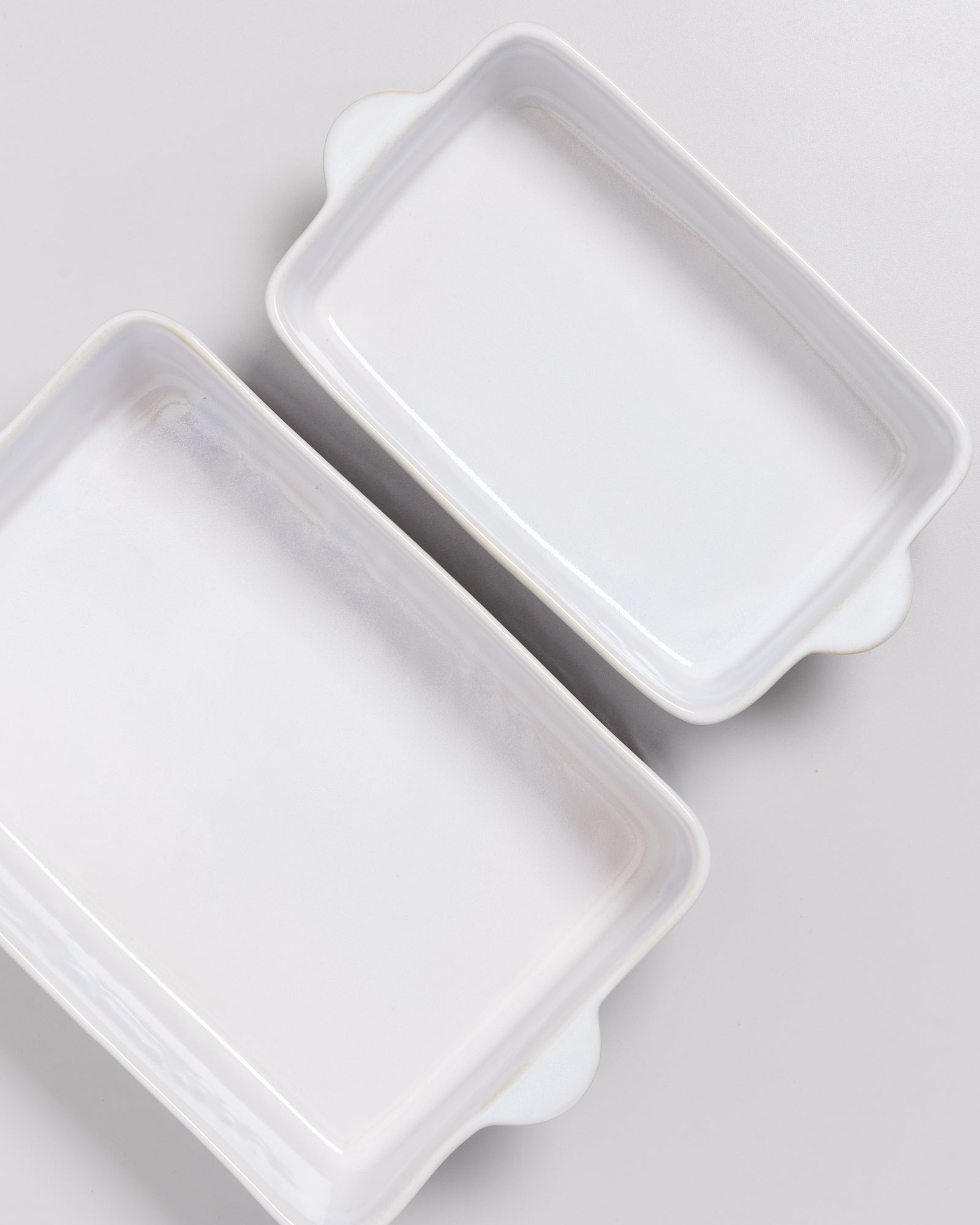 Zwei weiße rechteckige Keramik-Auflaufformen mit Griffen auf einer grauen Oberfläche.
