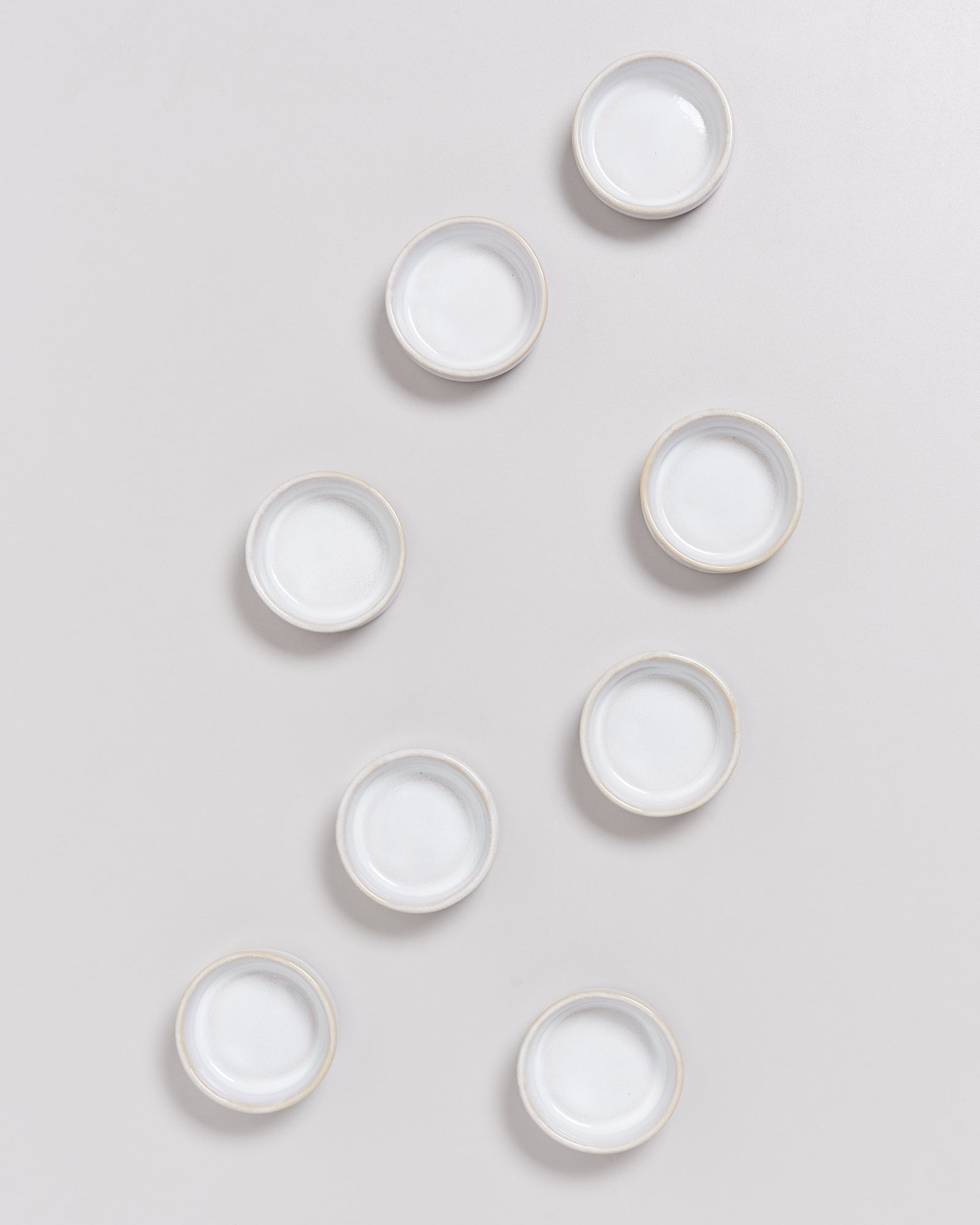 Acht kleine, weiße, runde Schälchen liegen auf einem hellgrauen Hintergrund verstreut.