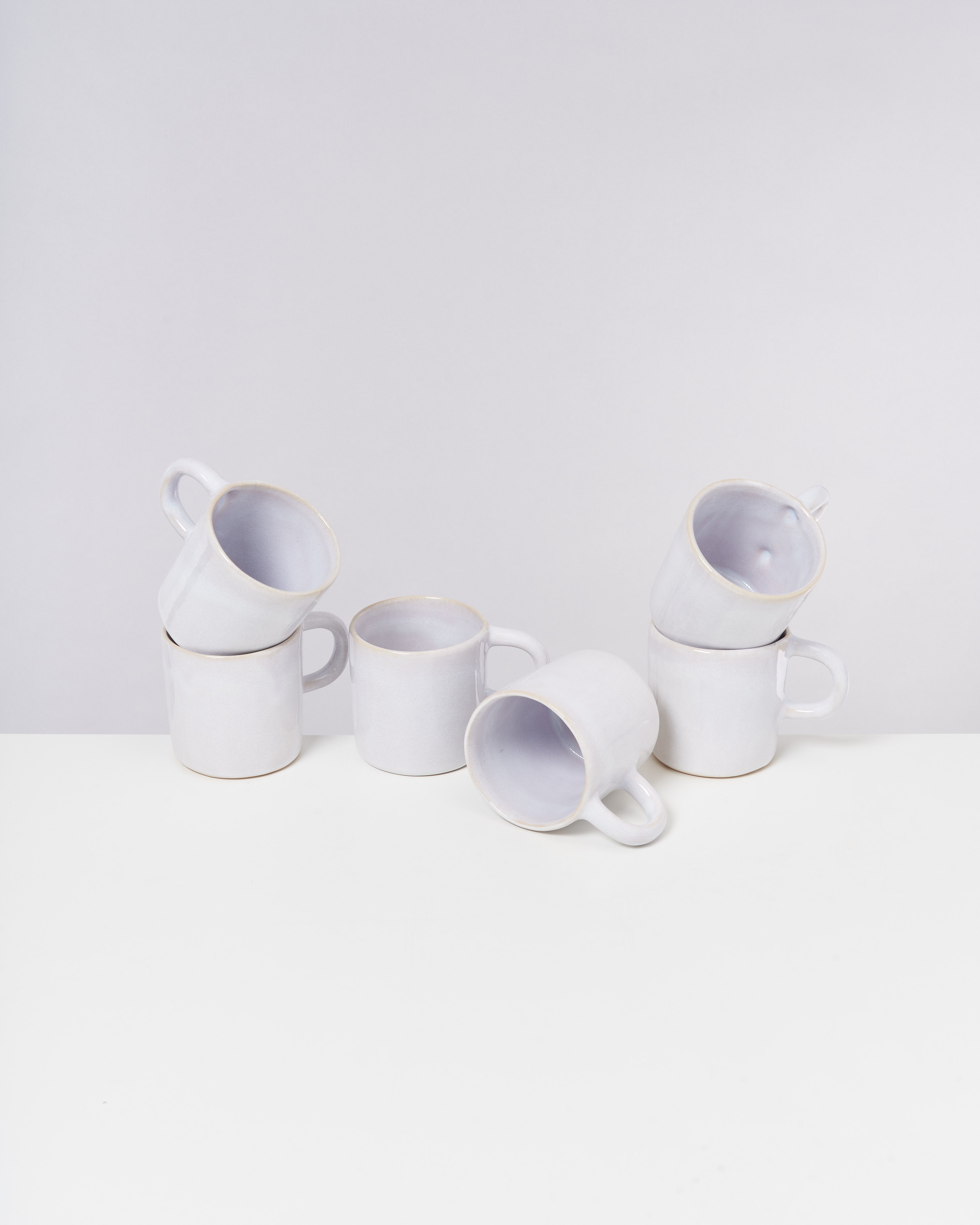 Sechs weiße Tassen stapeln sich ungleichmäßig auf einem weißen Tisch vor einem grauen Hintergrund.
