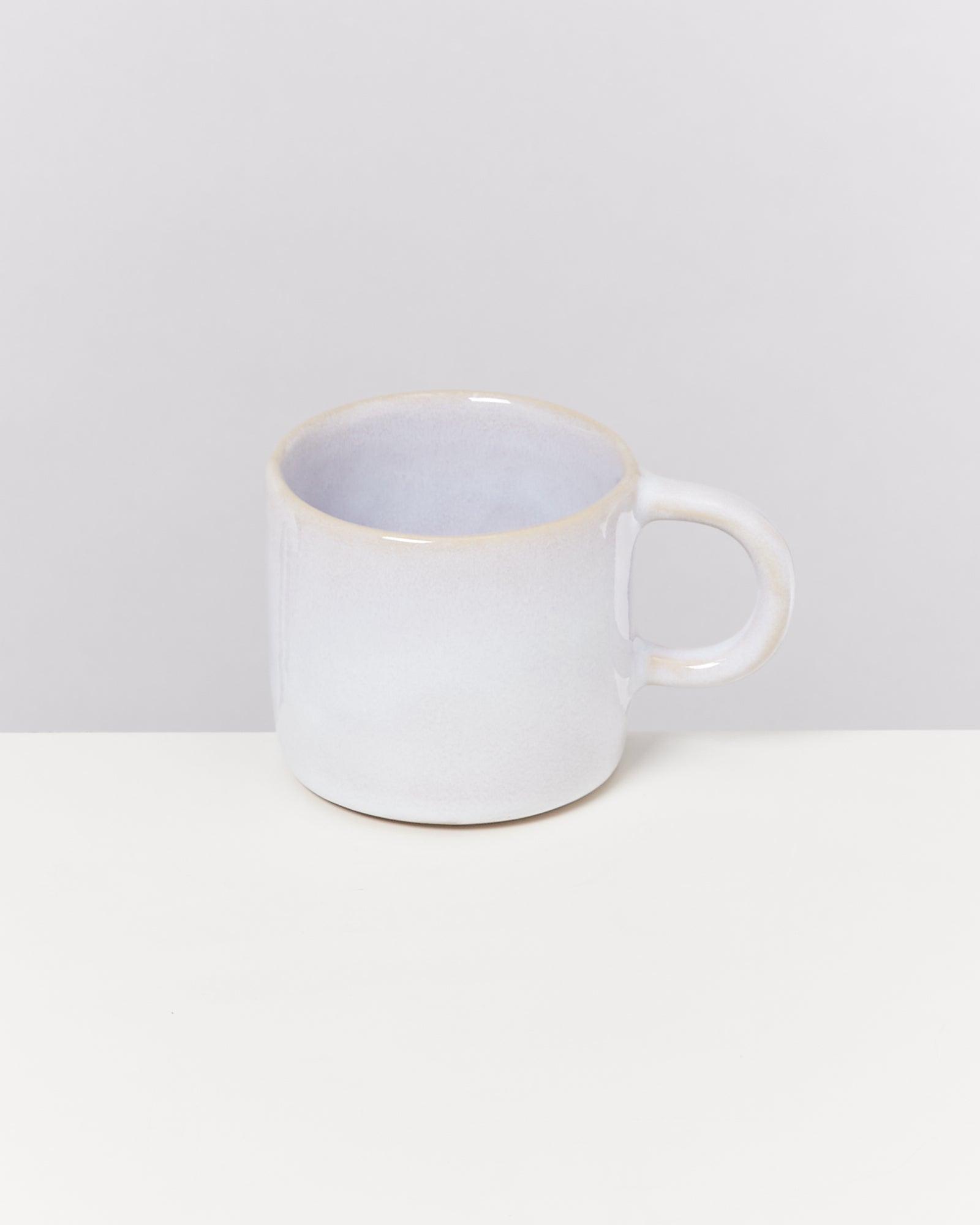 Weiße Keramiktasse mit Henkel steht auf einem weißen Tisch vor grauem Hintergrund.