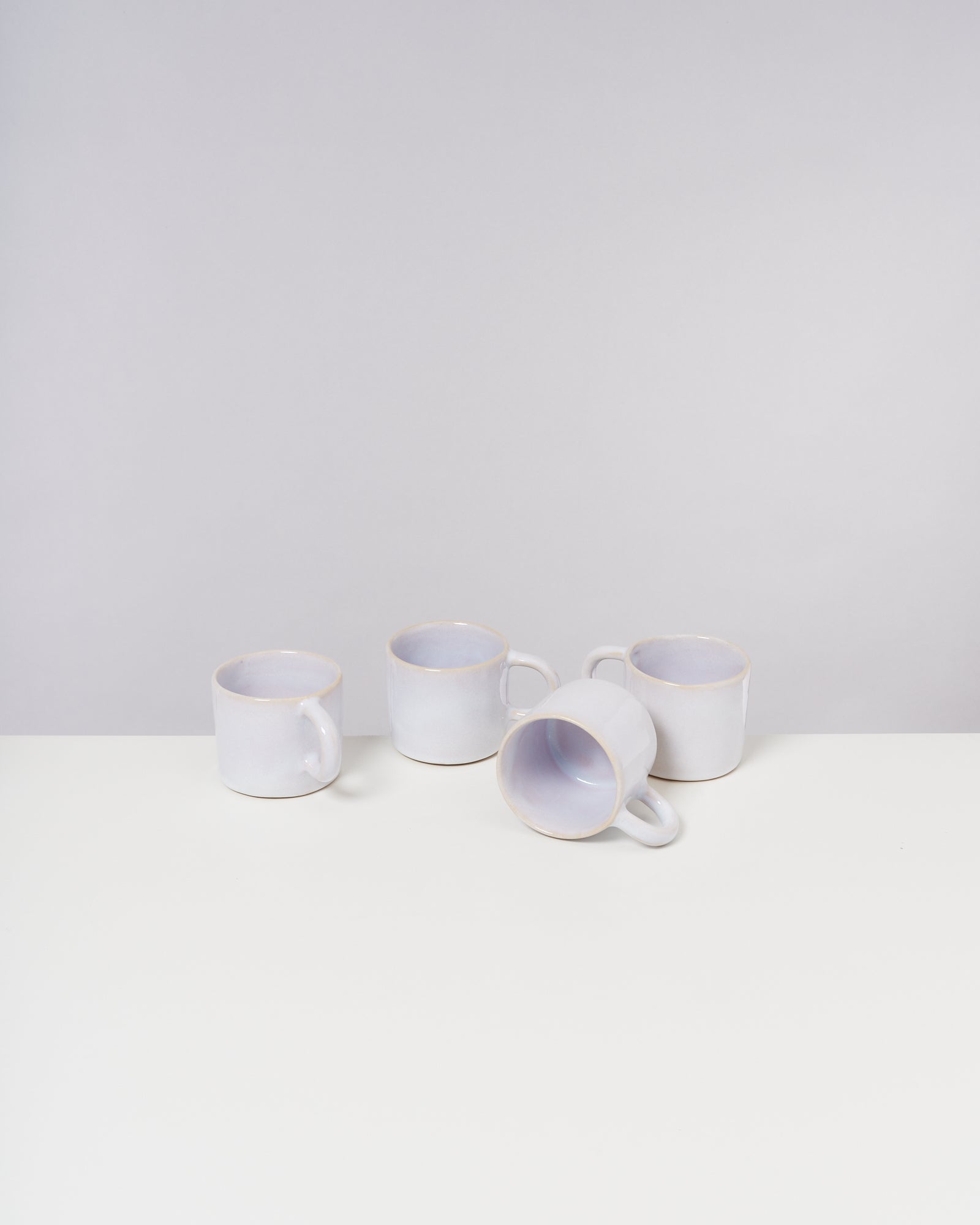 Vier weiße Tassen, eine liegend, auf einem weißen Tisch vor grauem Hintergrund.