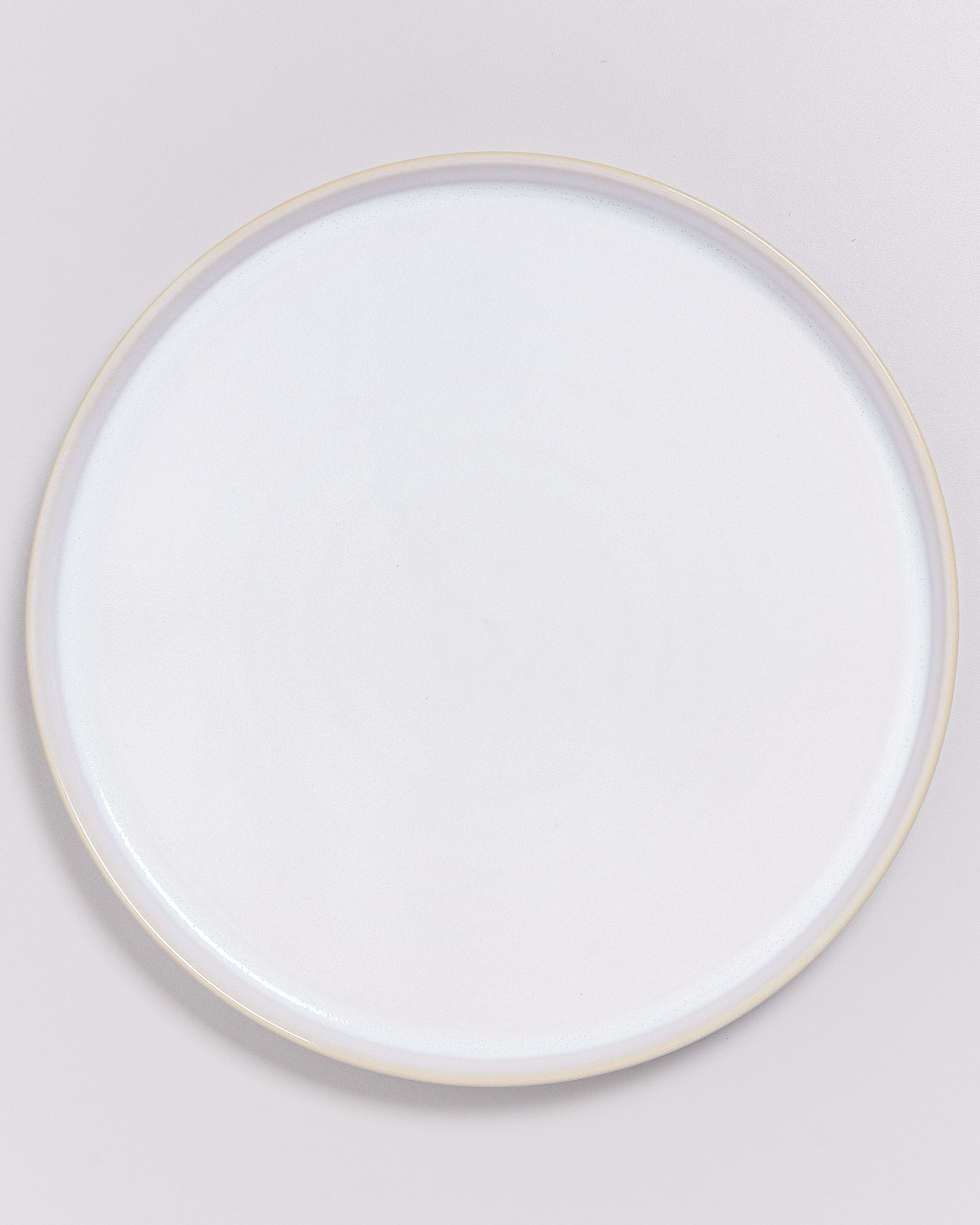 Ein einfacher, weißer, runder Teller mit einem schmalen, hellbraunen Rand auf einem grauen Hintergrund.