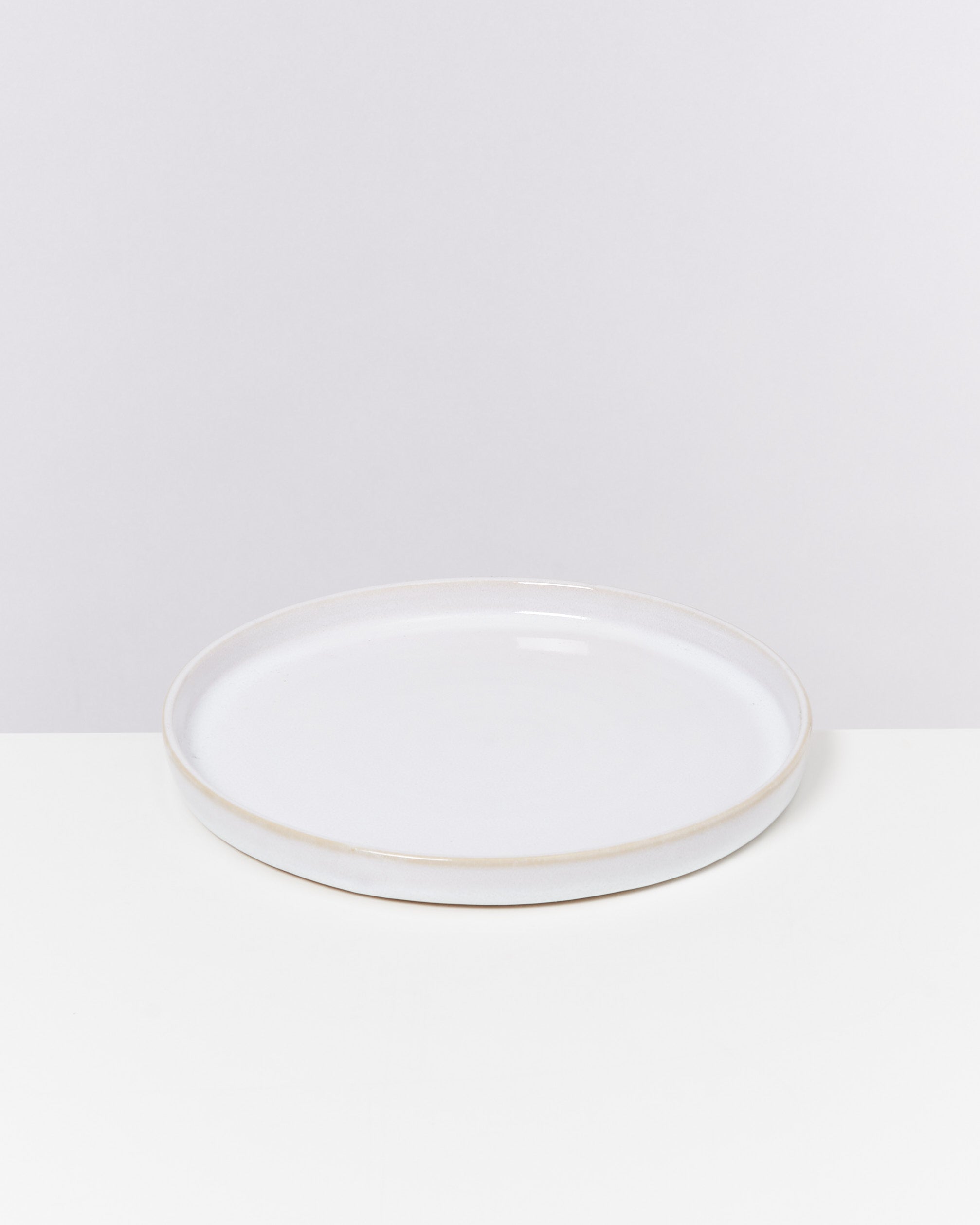 Ein flacher, weißer Keramikteller auf einer weißen Oberfläche vor einem hellgrauen Hintergrund.