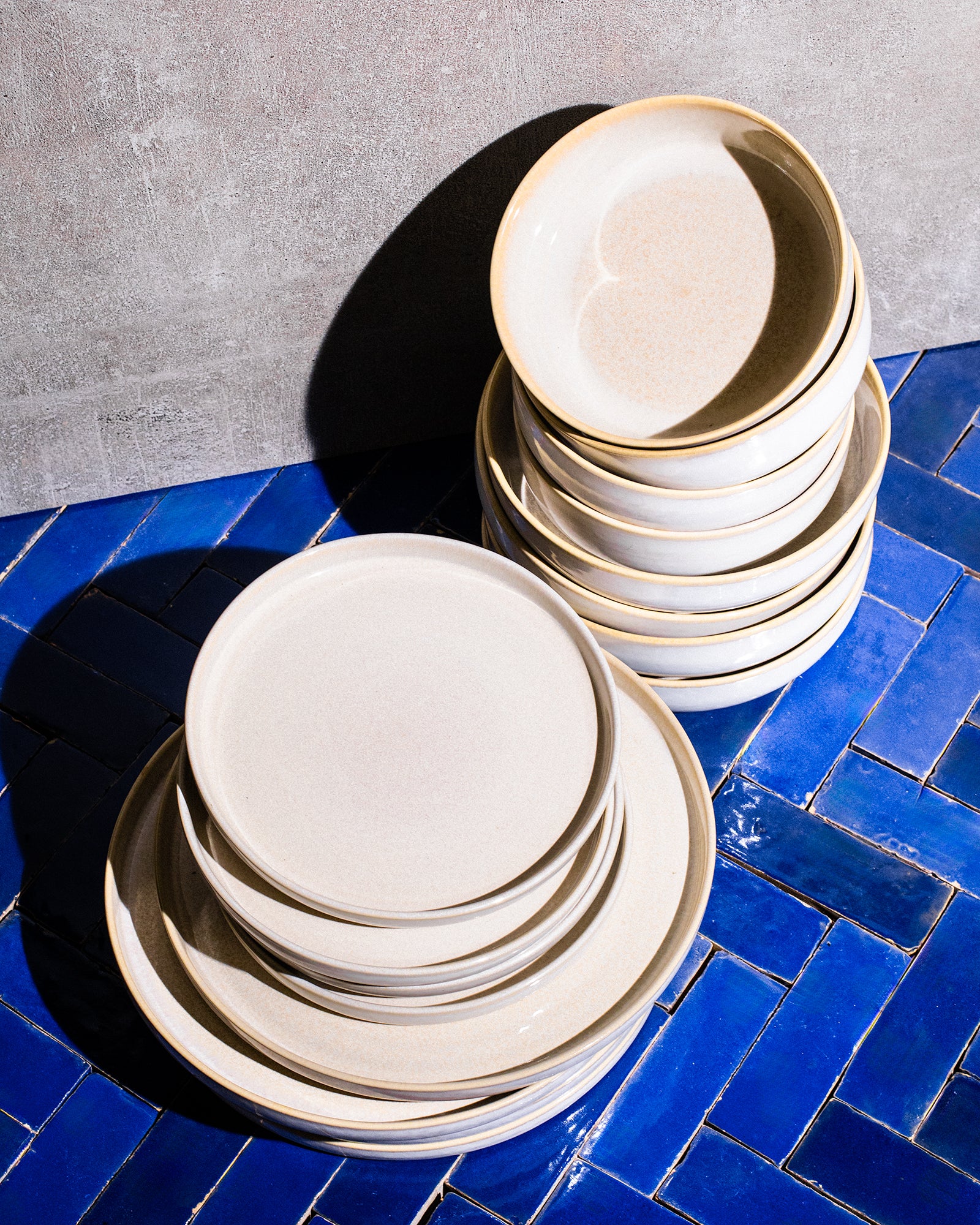 Gestapelte weiße Keramikteller auf blauen Fliesen vor einer grauen Wand.