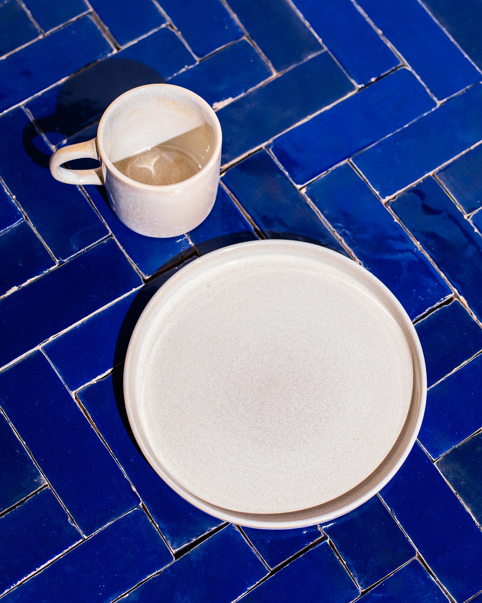 Beige Tasse und Teller auf einem blauen, gefliesten Tisch in hellem Sonnenlicht.