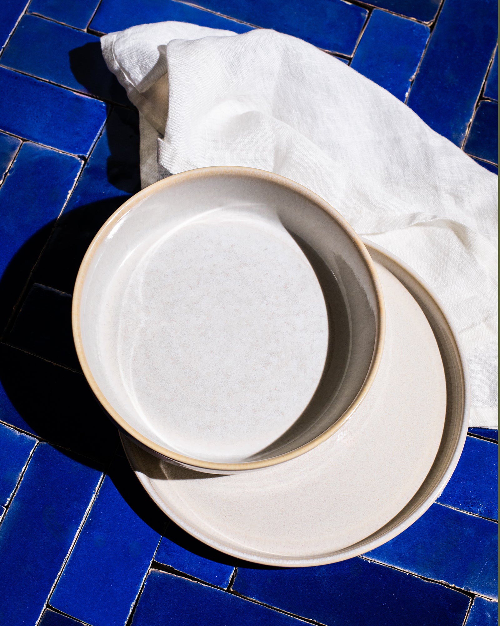 Zwei beige Teller auf blauen Fliesen, umgeben von einem weißen Stofftuch.